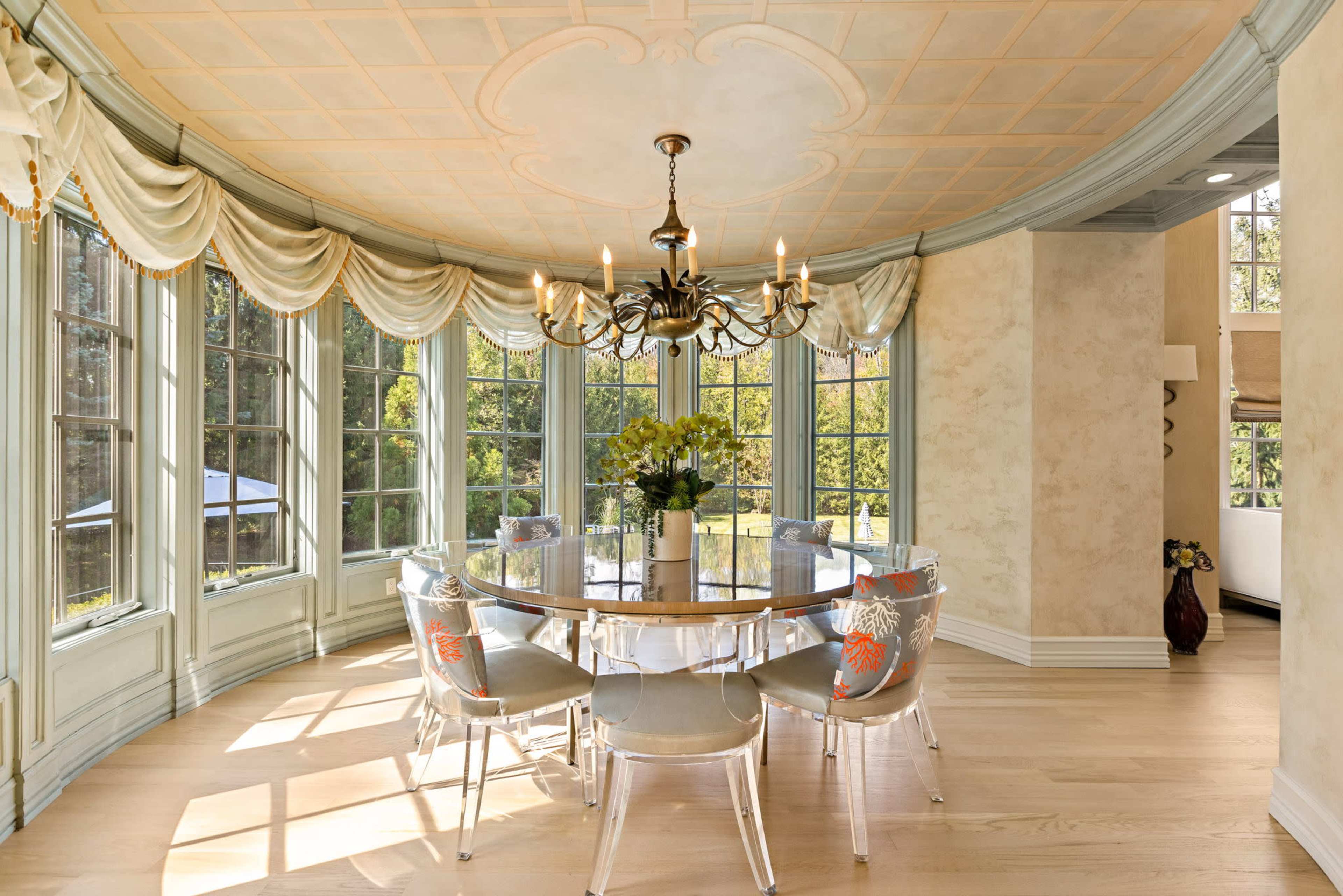 A round dining table with transparent chairs is set in a sunlit room featuring large windows and soft, draped curtains.