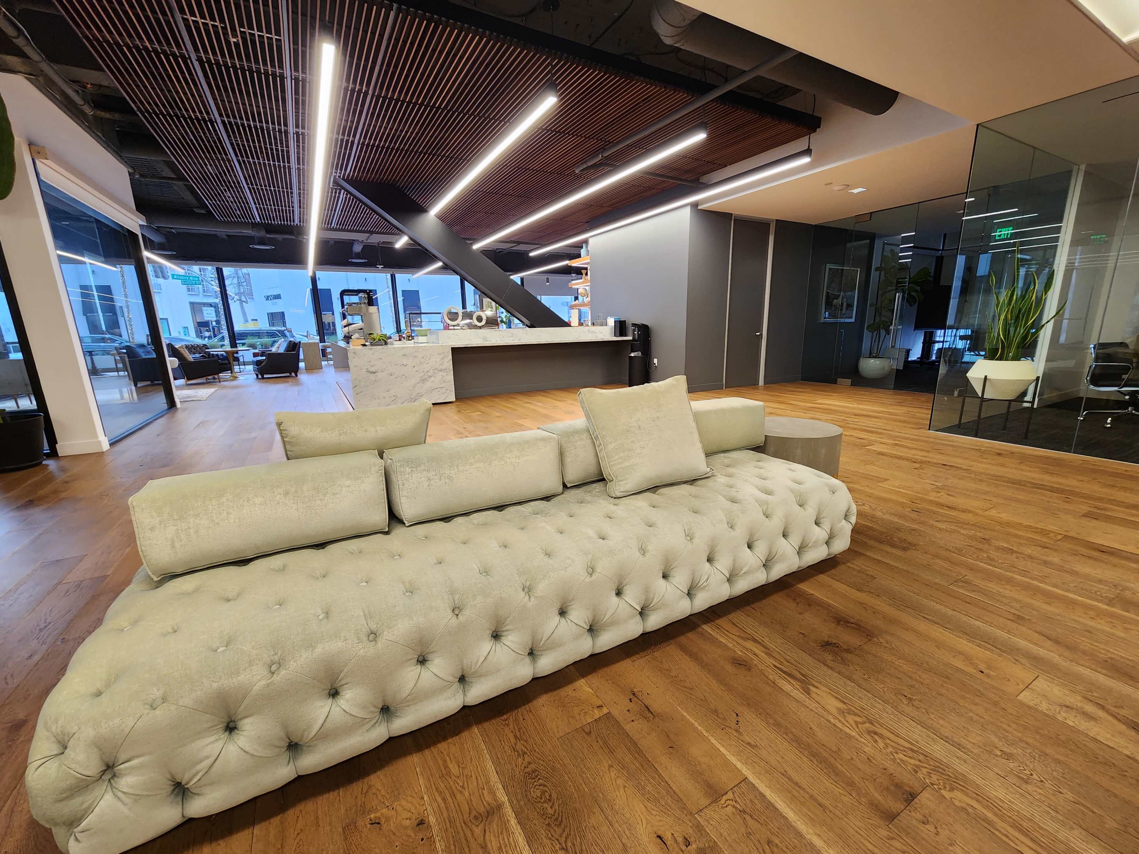 A modern office interior featuring a tufted light-colored sofa, wooden flooring, and an open layout with glass partitions.