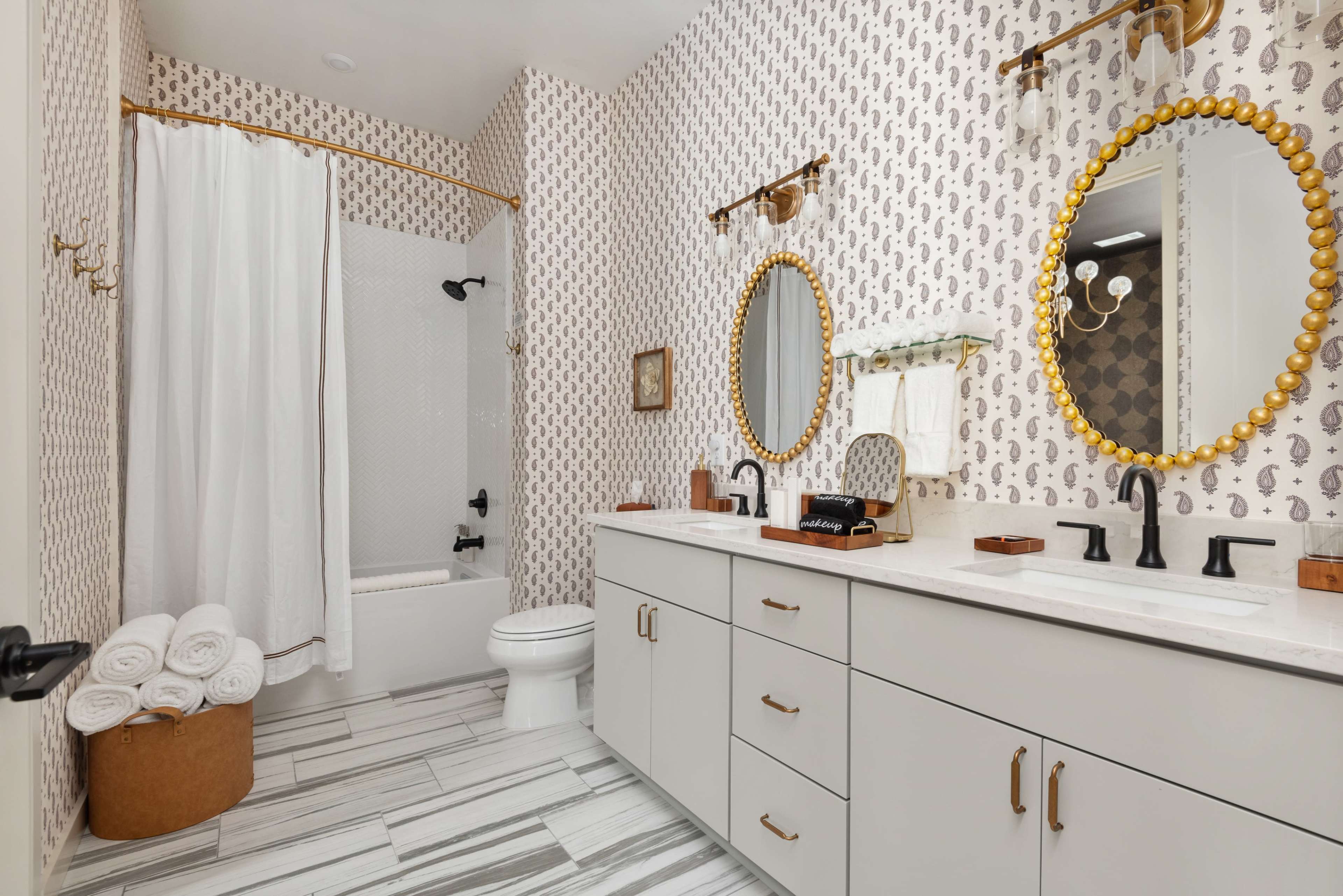 The bathroom features a double vanity with round mirrors, a tub-shower combo, and neatly stacked towels beside a decorative basket.