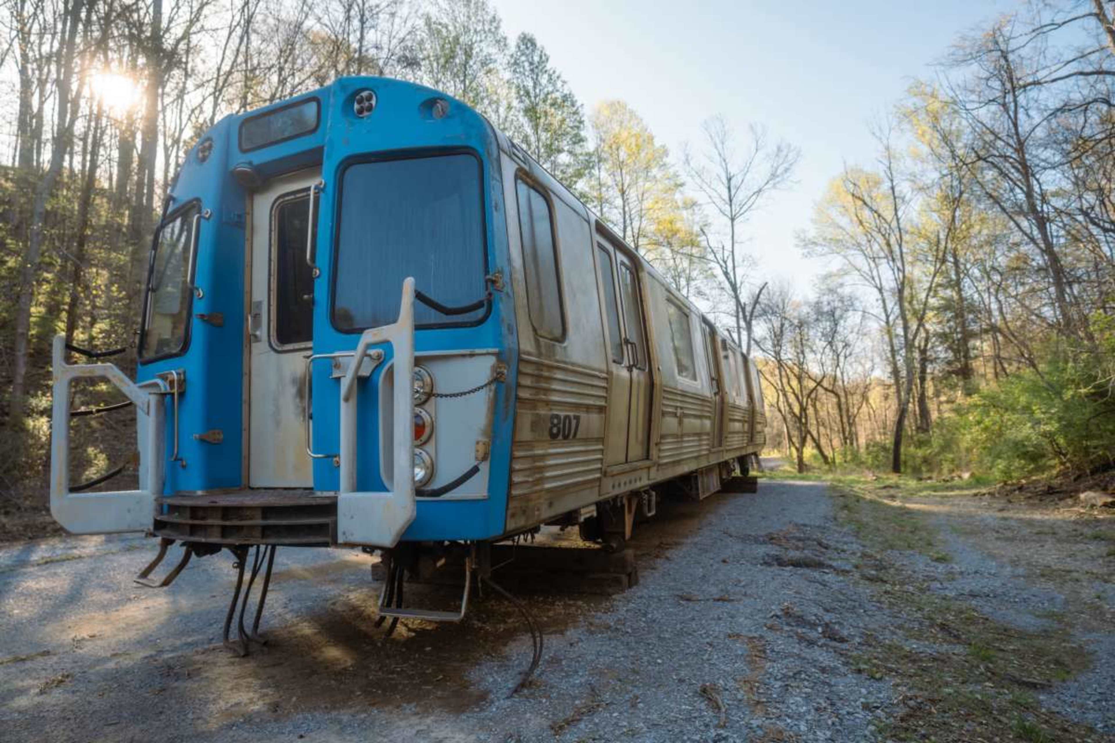 A weathered blue and silver train car sits on a gravel path surrounded by trees.