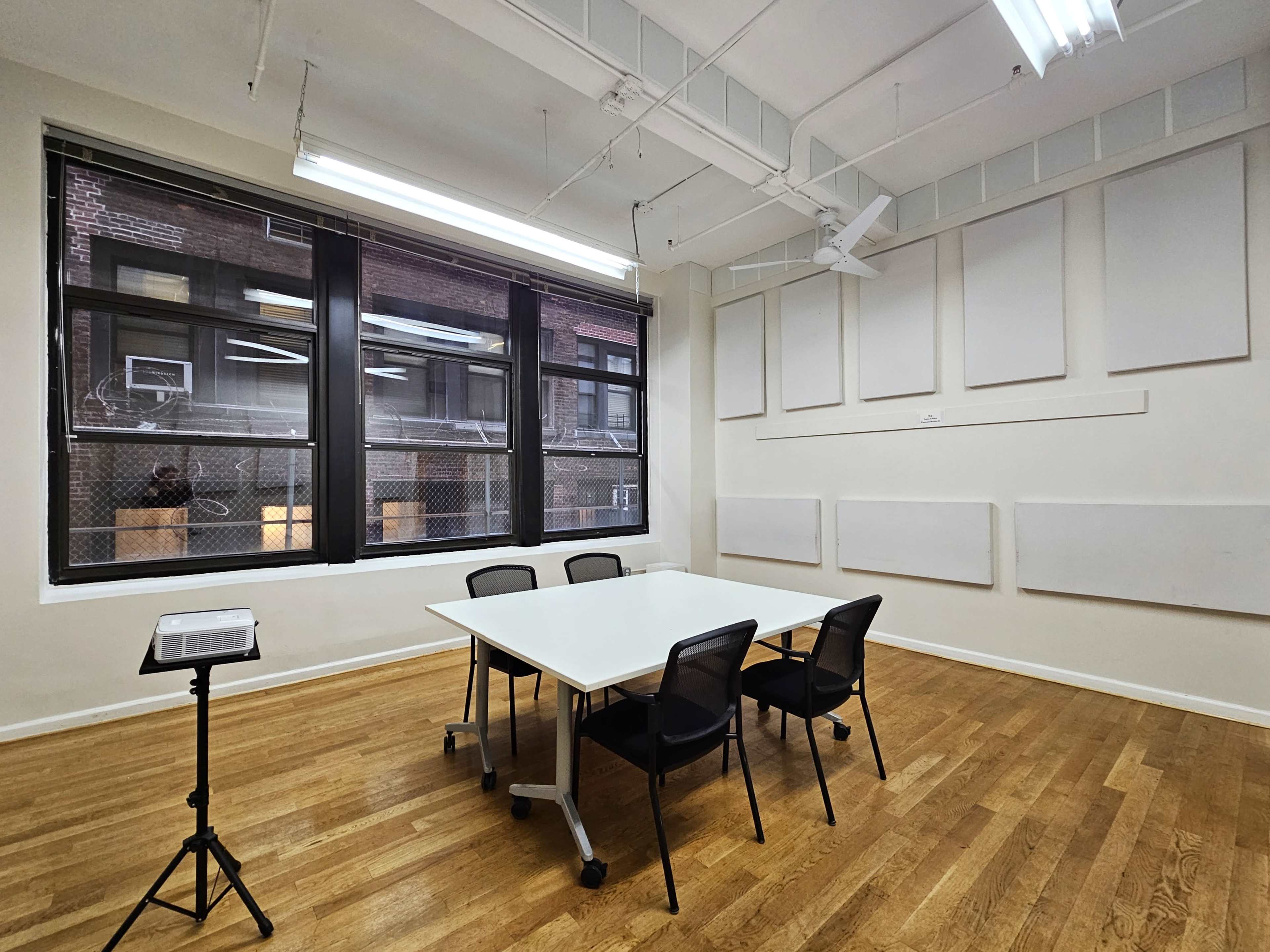 A conference room features a white table surrounded by four chairs, with large windows showing a view of the outside and empty wall panels.