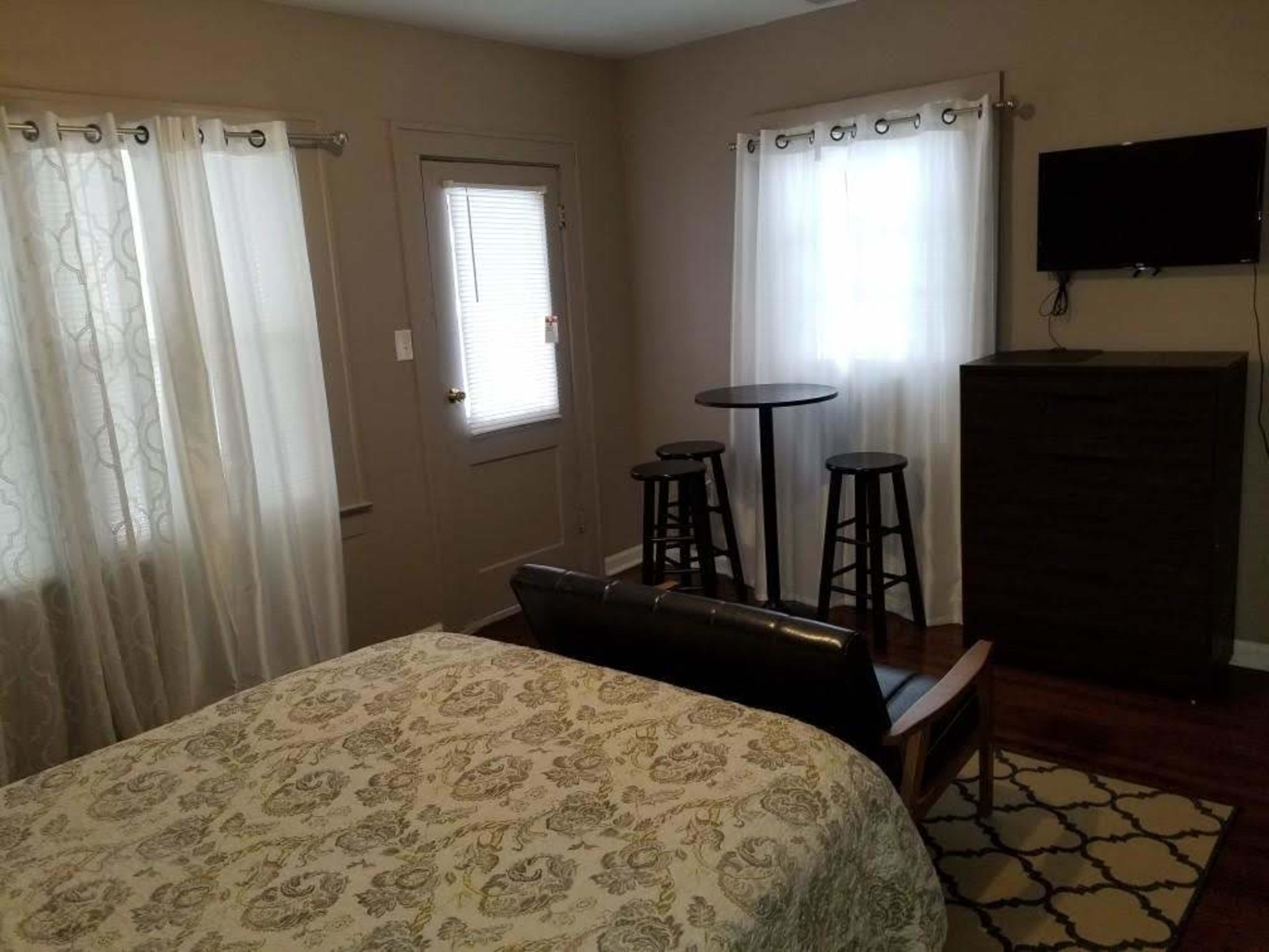 A small bedroom with a bed, a set of stools around a tall table, a dresser, and windows with white curtains.
