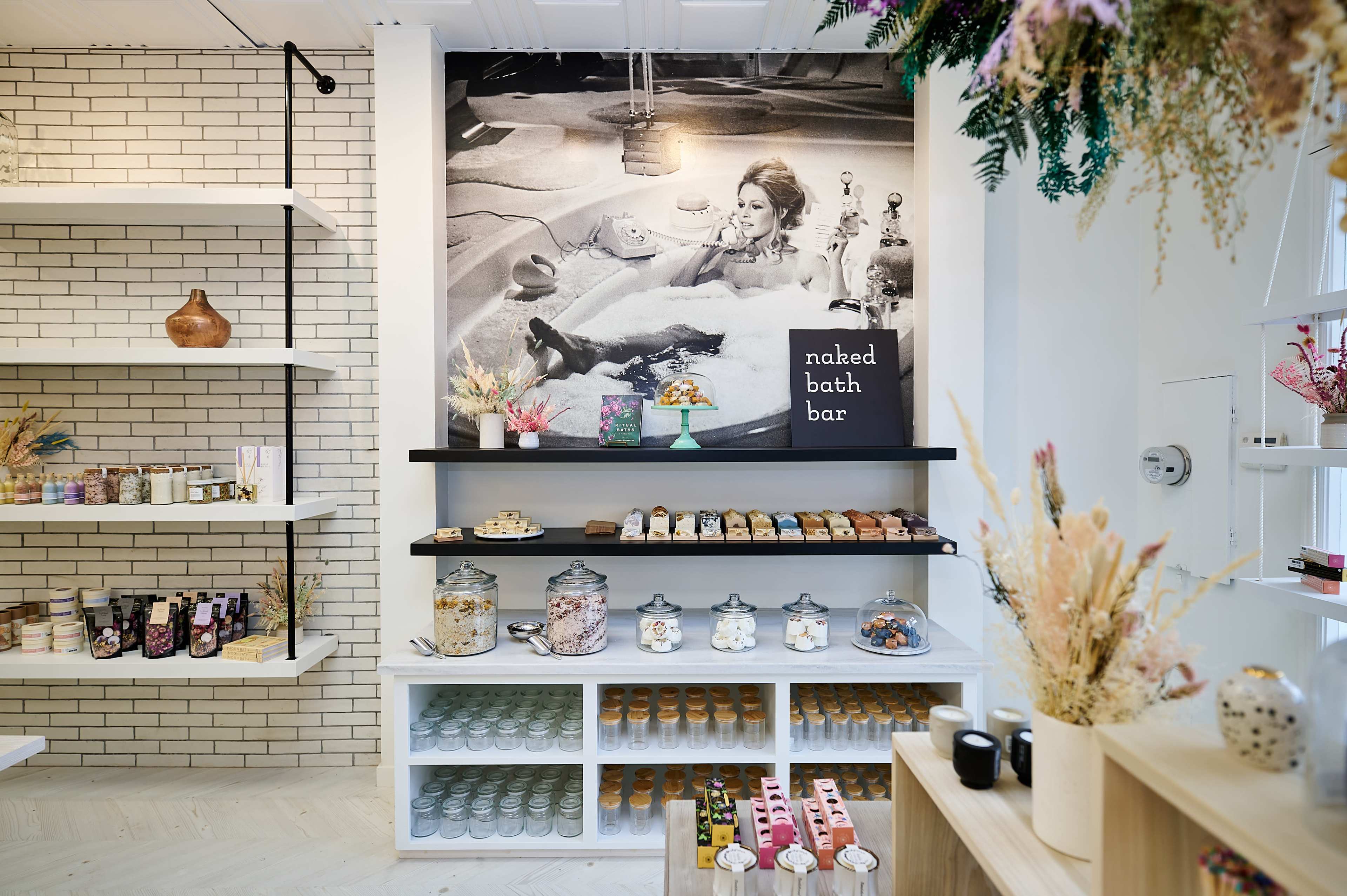 A stylish bath product store with shelves displaying various jars and packaged goods, featuring a large black-and-white photo of a woman in a bathtub on the back wall.