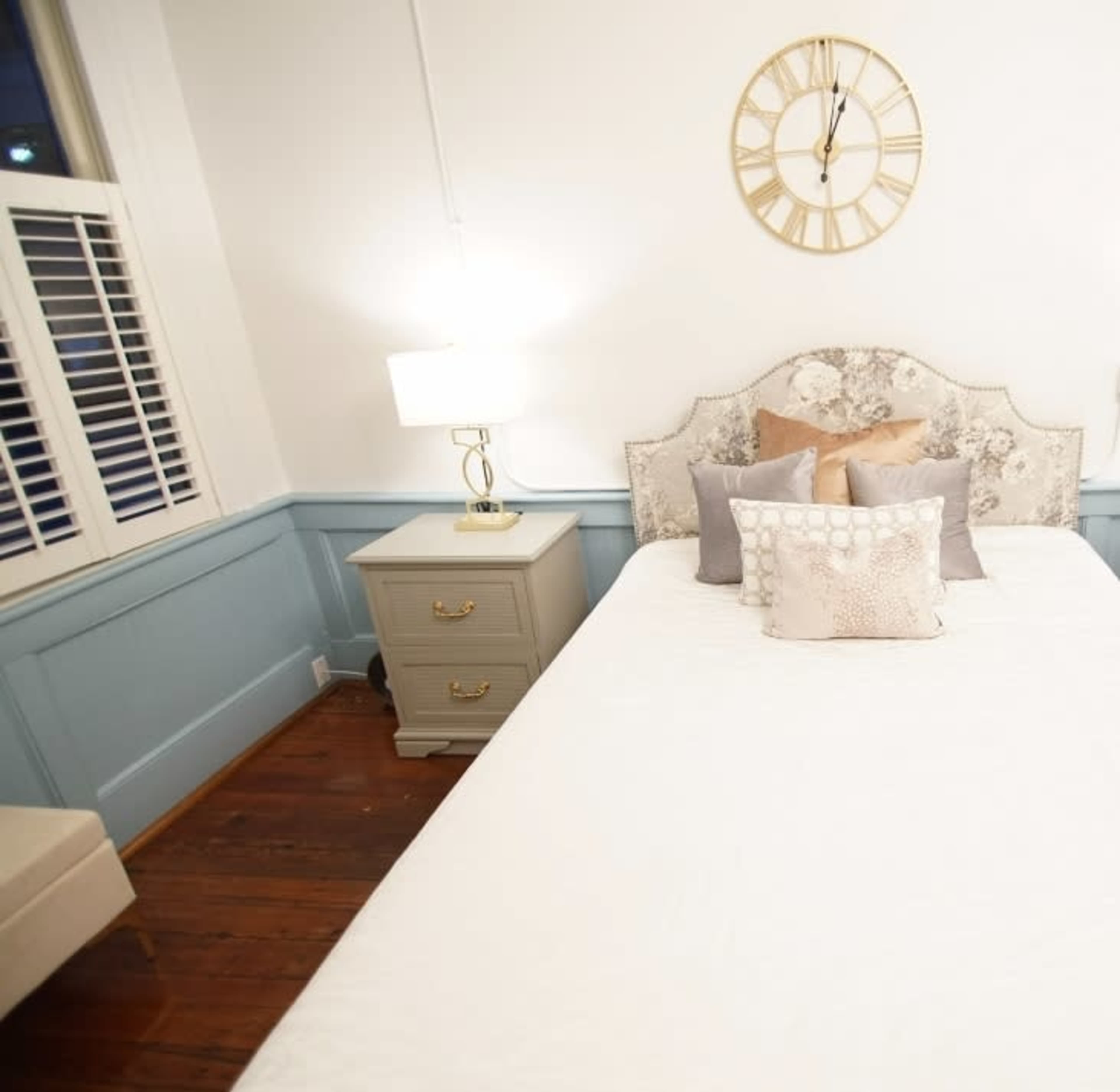 A neatly made bed is positioned in a well-lit room with light blue paneling and a wall clock above the headboard.