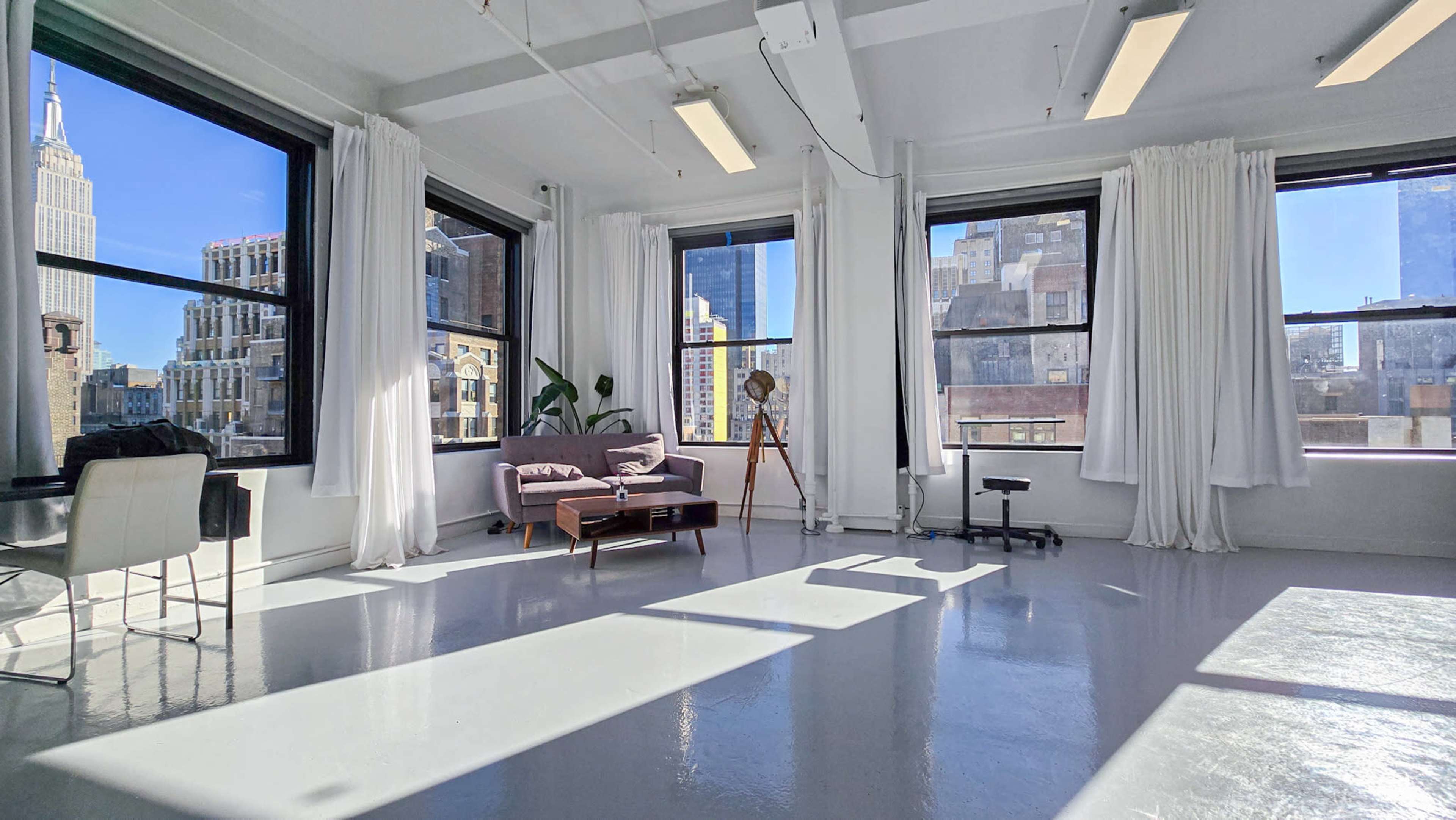 The image shows a bright, spacious room with large windows, featuring a purple sofa, a wooden tripod, and white curtains, set against an urban skyline.
