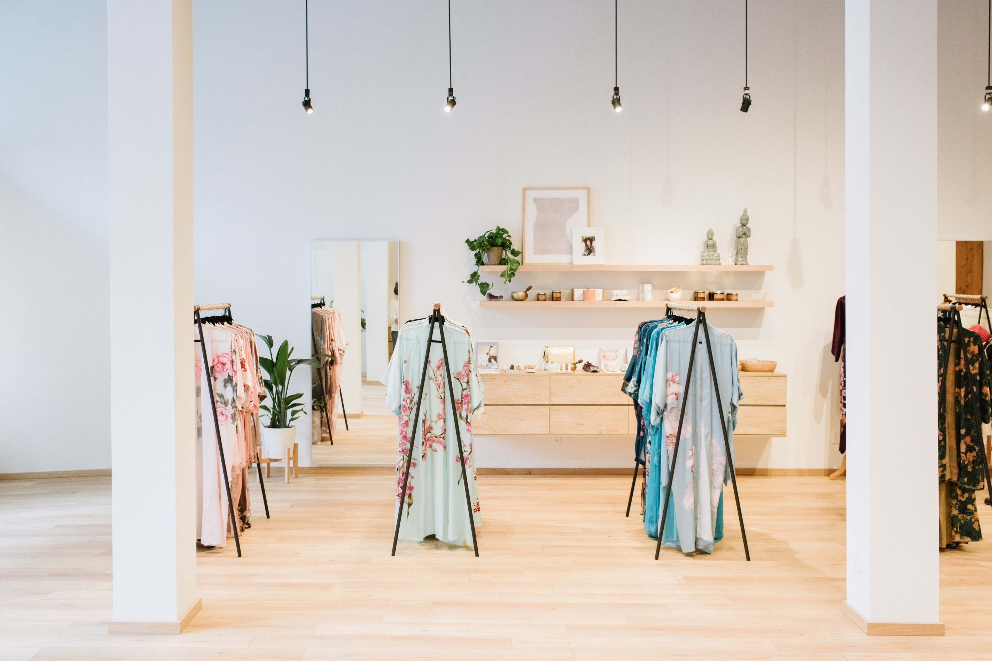 A clothing boutique with racks displaying garments, a wooden counter, a mirror, and decor plants against a light-colored interior.