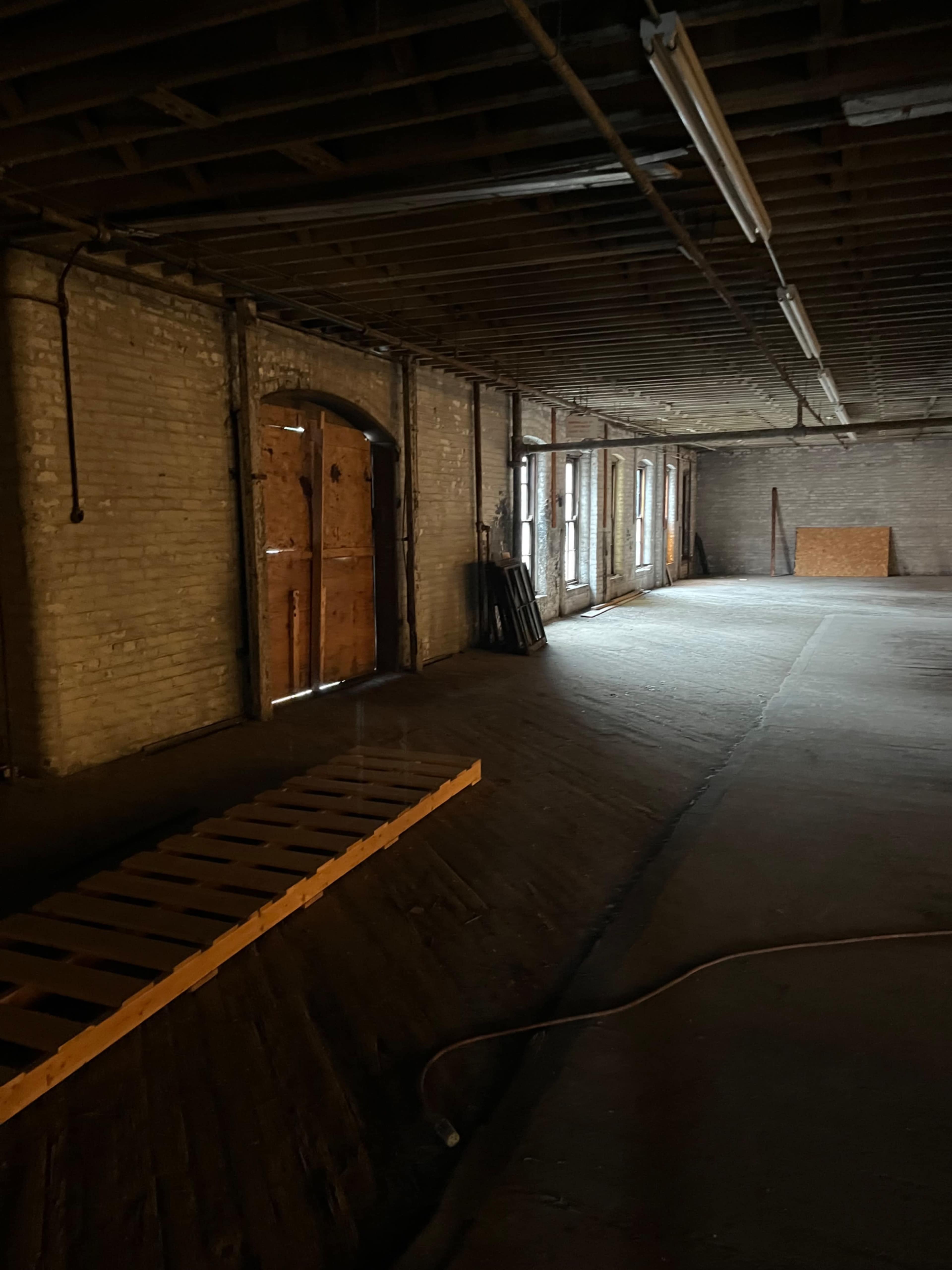 The image shows a dimly lit, empty industrial space with exposed brick walls, large windows, and a wooden ramp leading to an arched doorway.