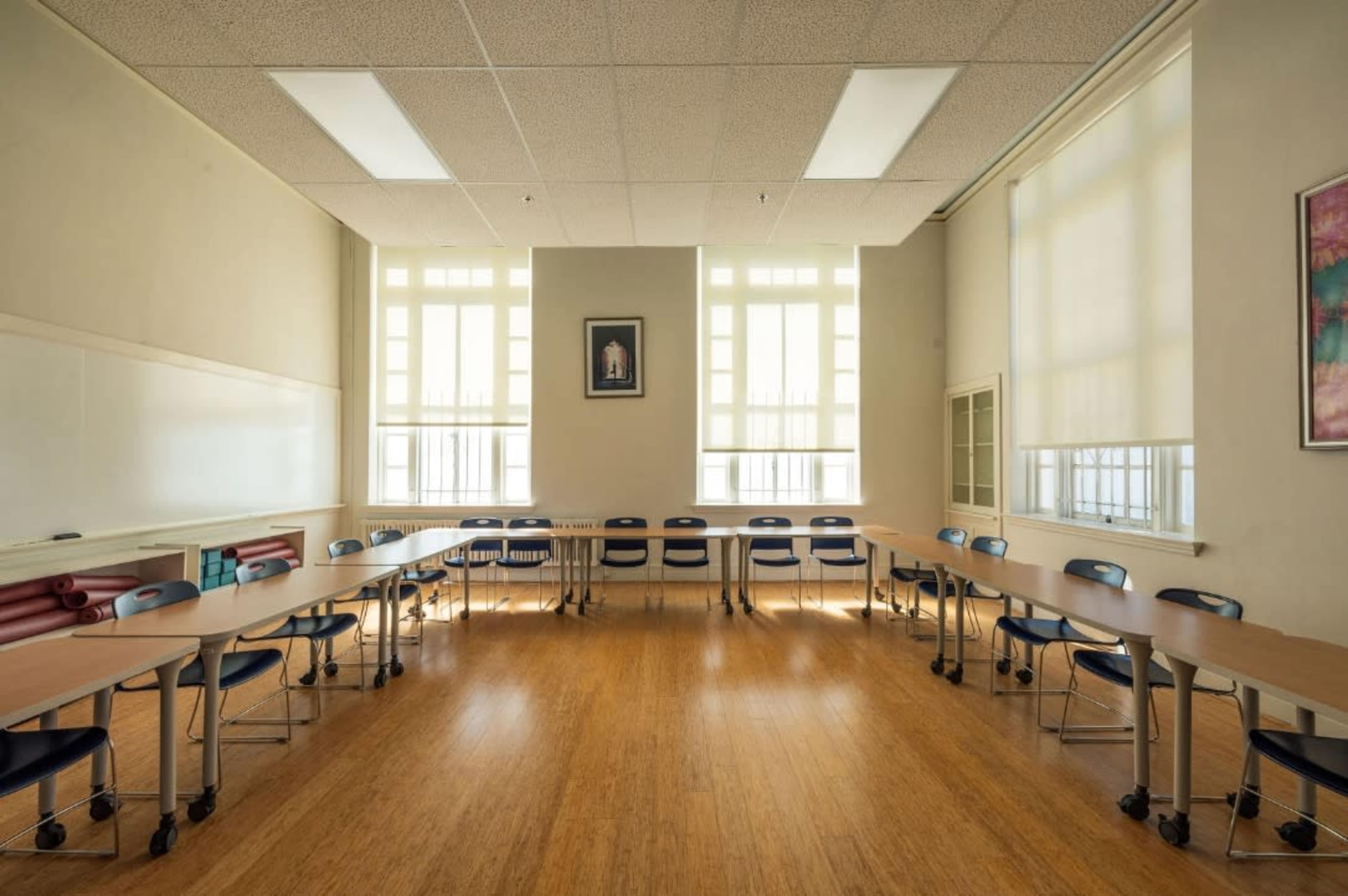 A spacious classroom is arranged with tables and chairs in a circular formation, illuminated by large windows.