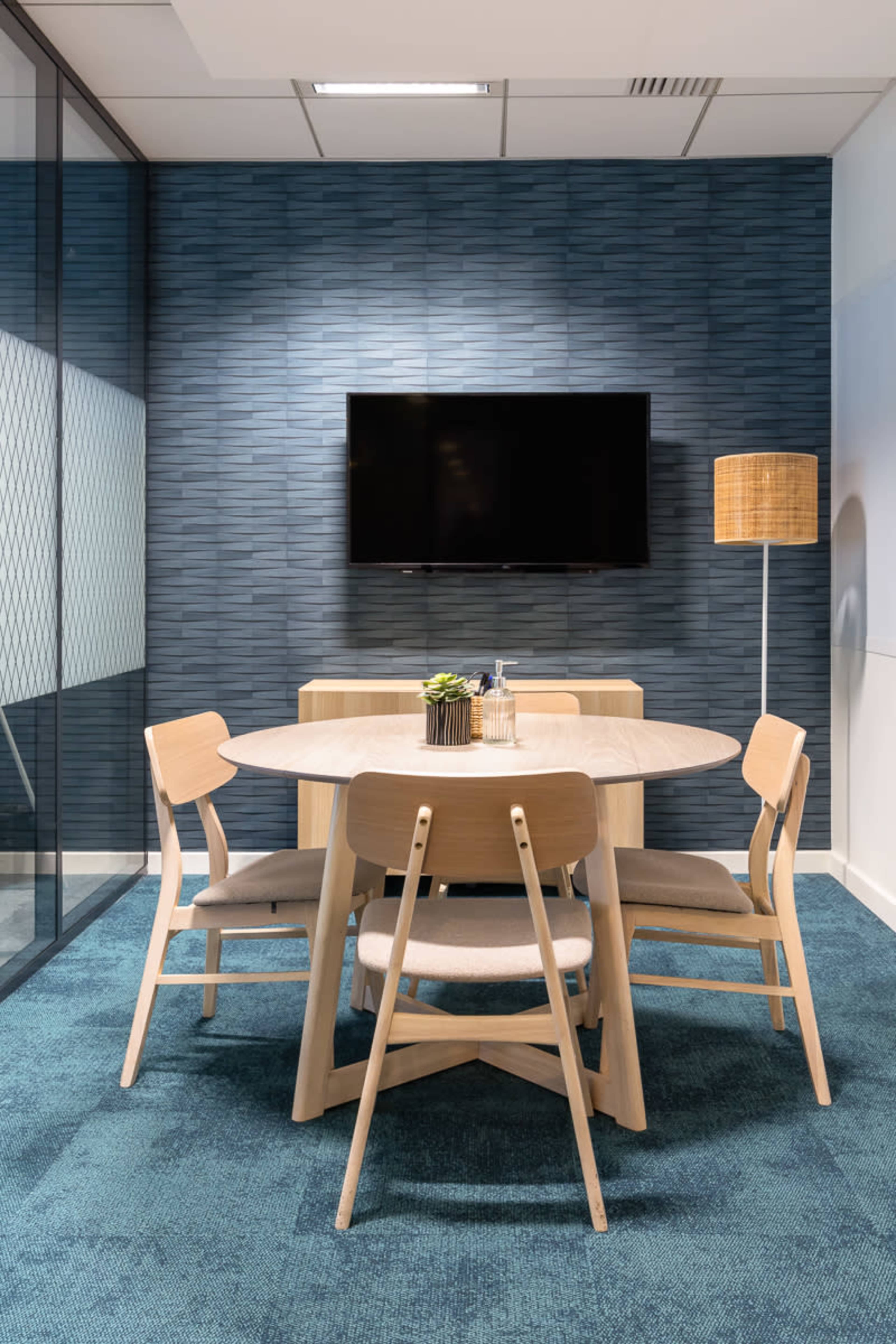 A modern meeting room features a round wooden table surrounded by four chairs, a television mounted on the wall, and a floor lamp, all set against a textured blue wall.