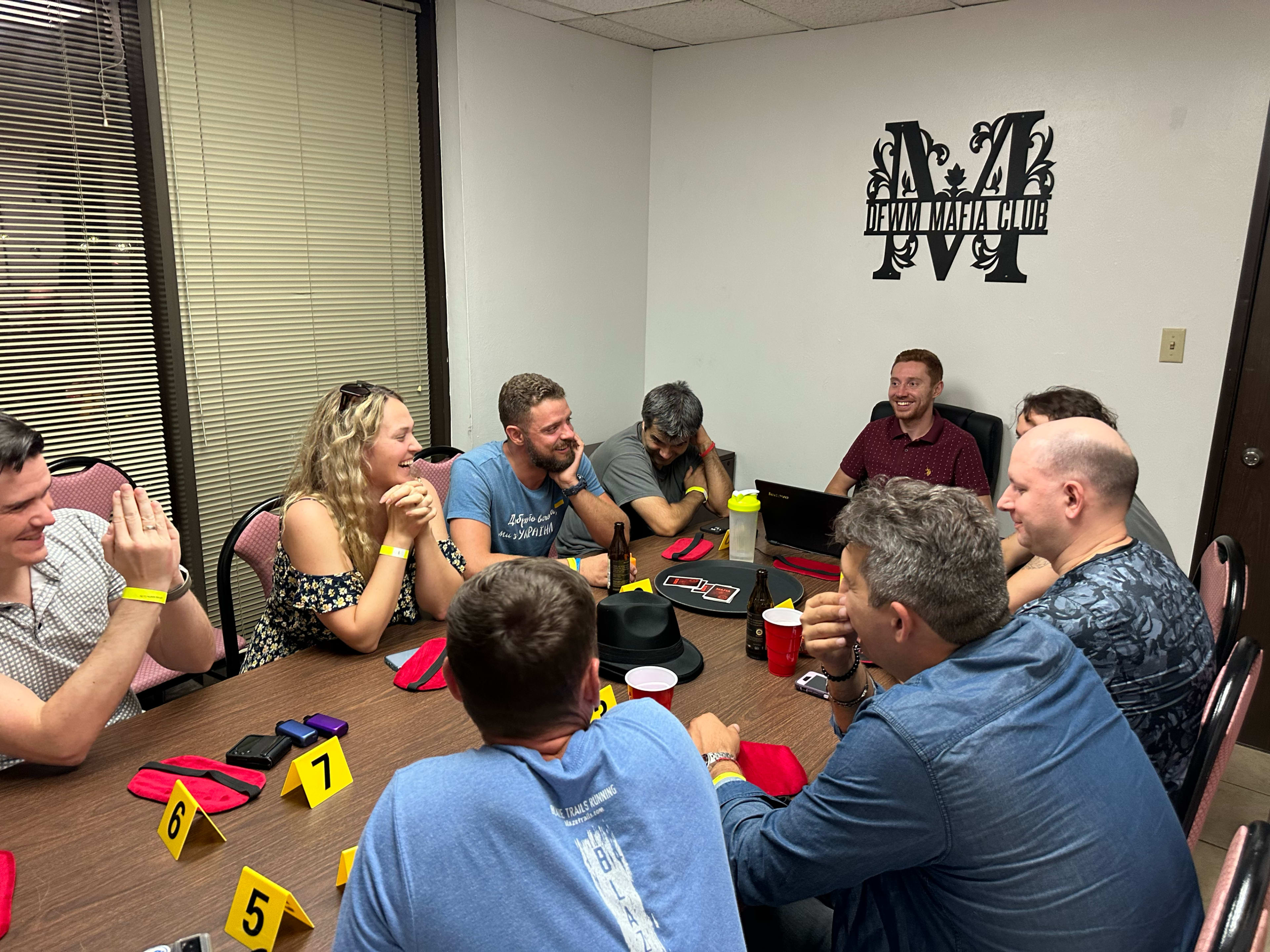 A group of people sits around a table in a room, engaged in conversation and activities, with numbered cards placed on the table.