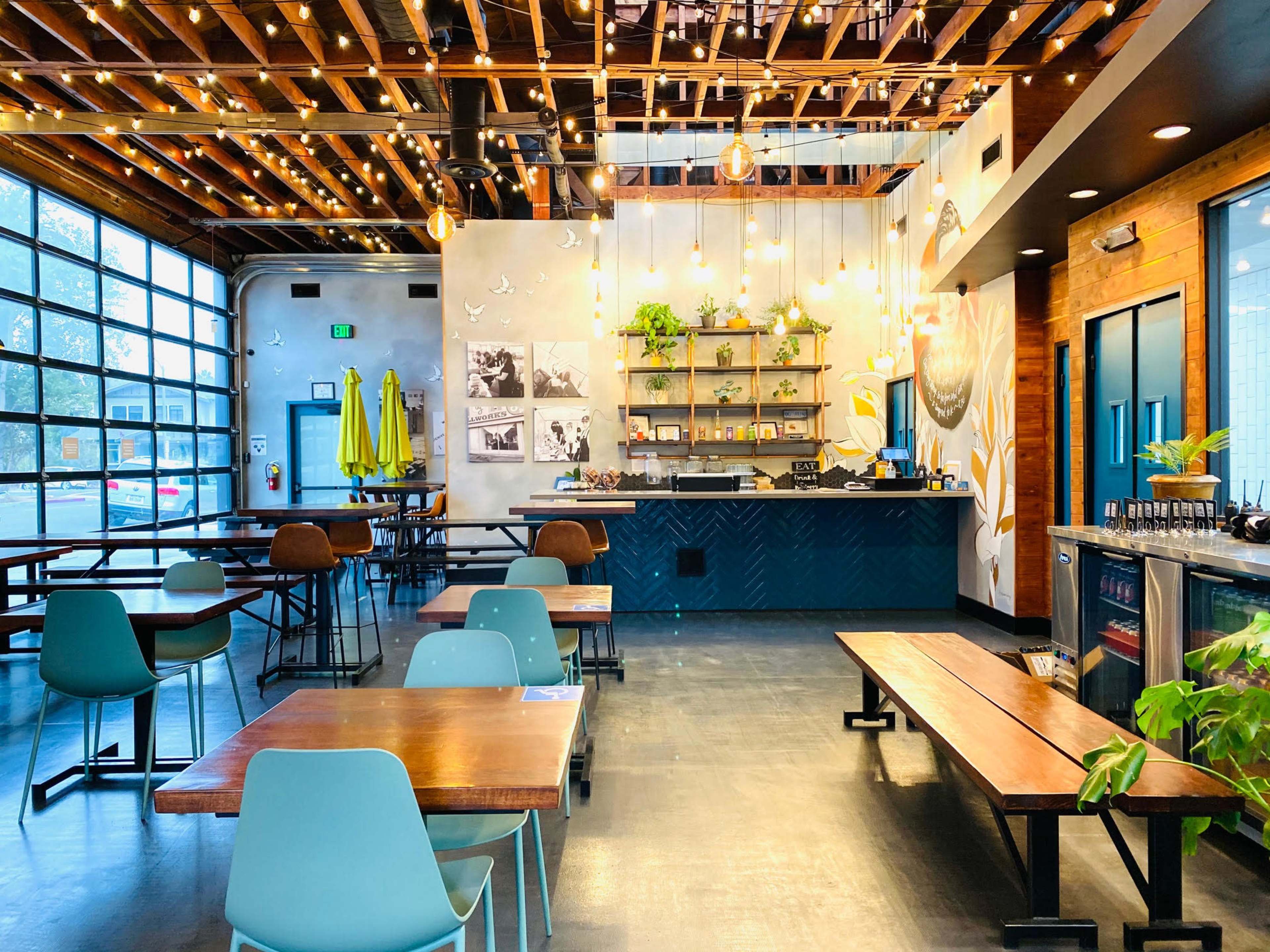 A modern café interior with wooden tables, teal chairs, and a counter adorned with plants and decorative lights.