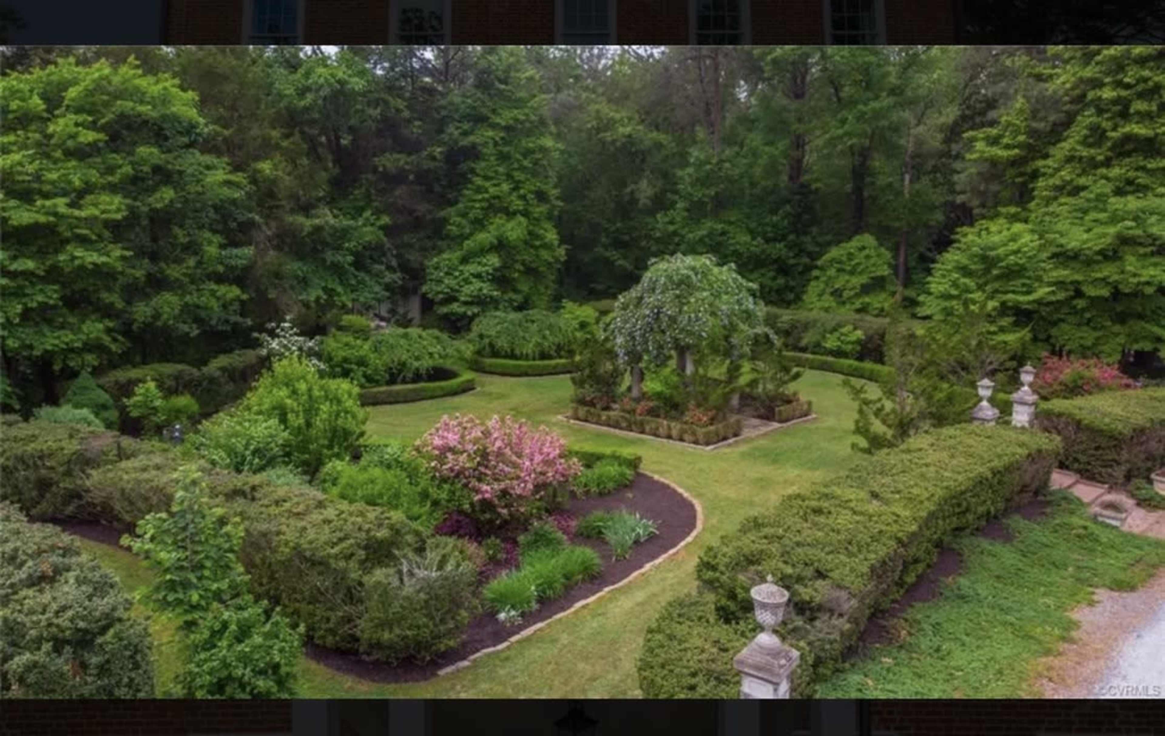 A well-maintained garden features neatly trimmed hedges, colorful flowerbeds, and a central tree surrounded by manicured paths.