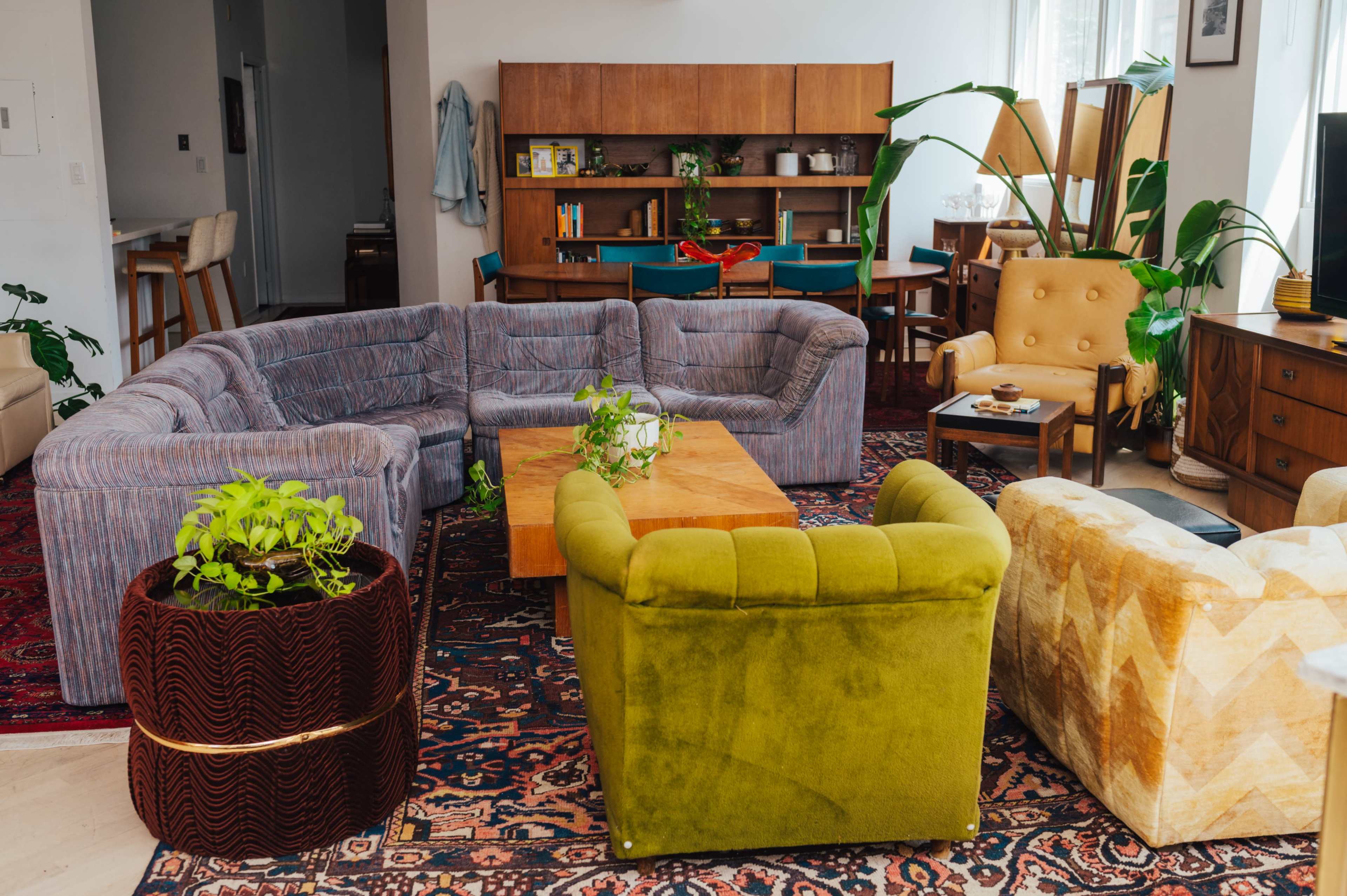 The image shows a modern living room with a sectional sofa, a green armchair, and a yellow armchair arranged around a wooden coffee table, surrounded by plants and mid-century furniture.