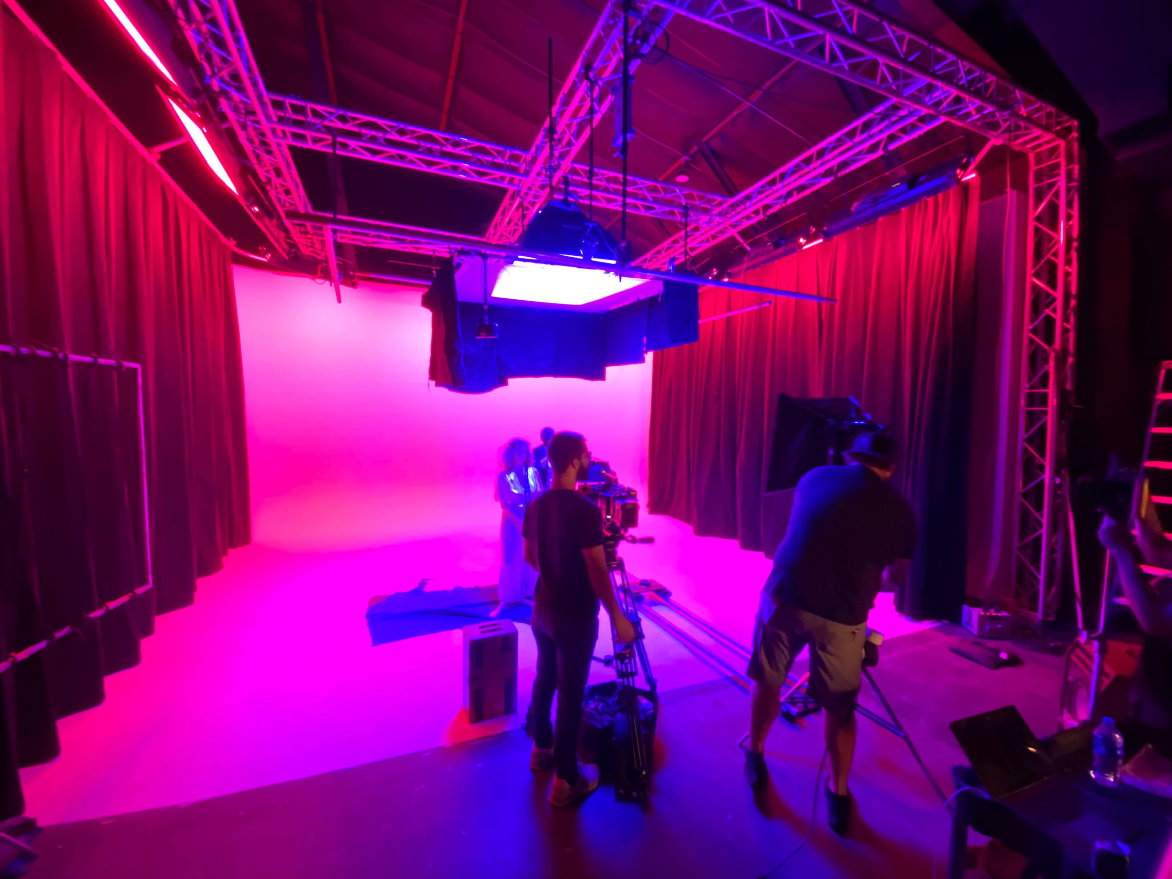 A film crew operates equipment in a brightly lit studio with pink and purple background lighting.