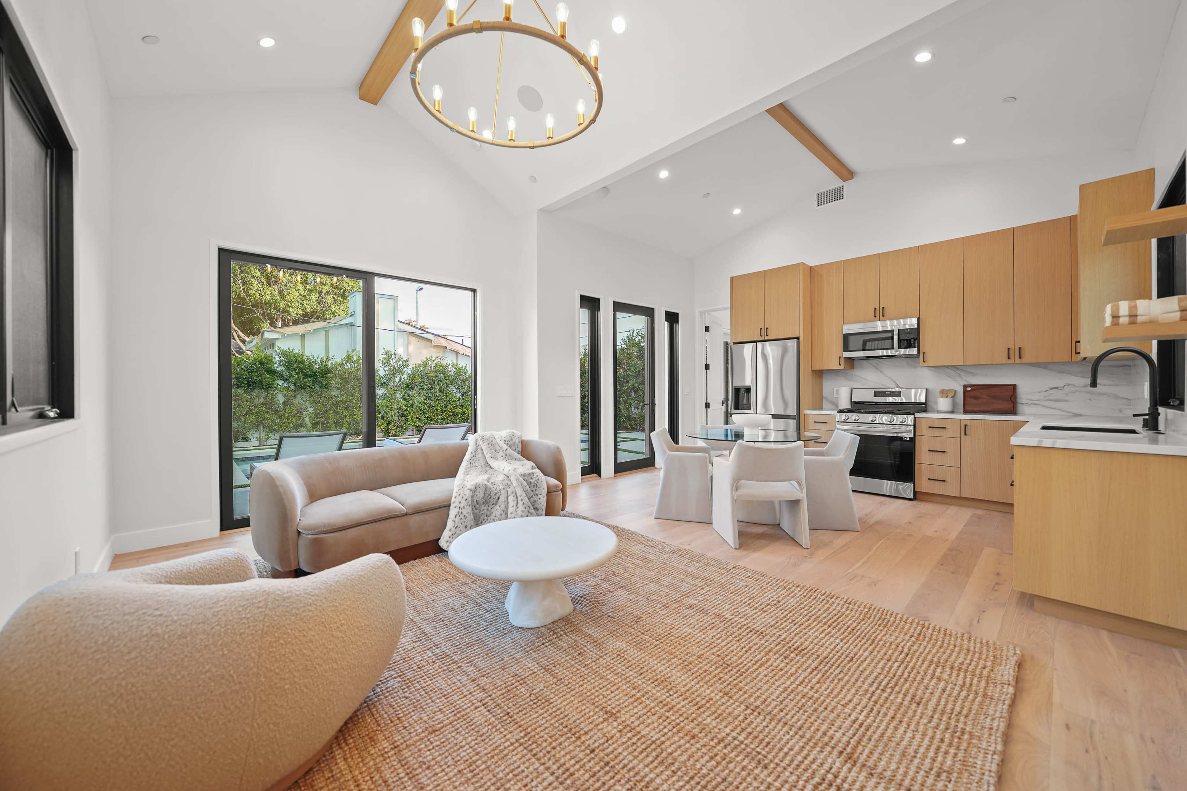 The image depicts a modern living room with a light-colored sofa, a round coffee table, and a kitchen area featuring wooden cabinetry and stainless steel appliances.
