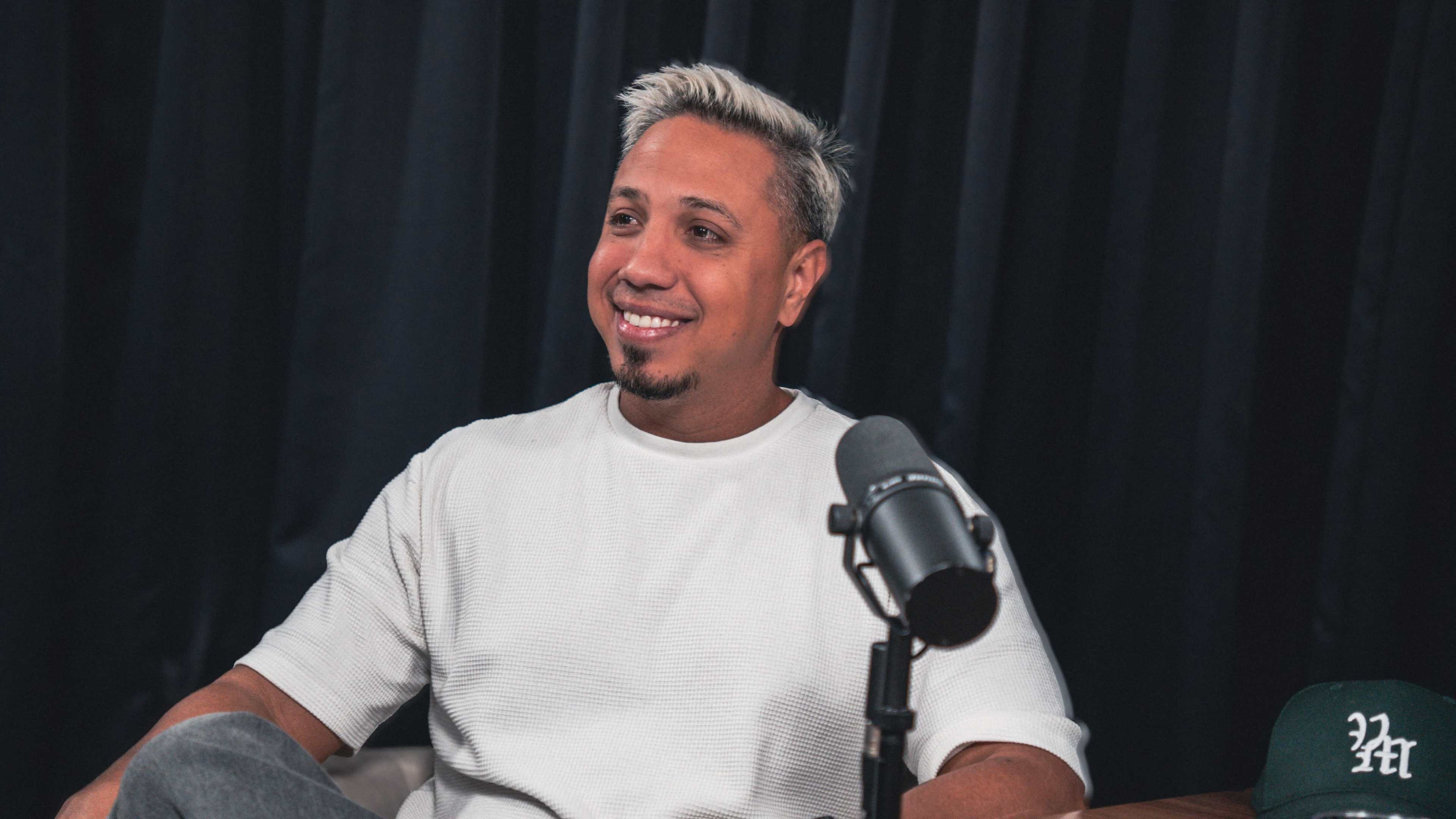 A man with light-colored hair smiles while sitting in front of a microphone in a recording studio.