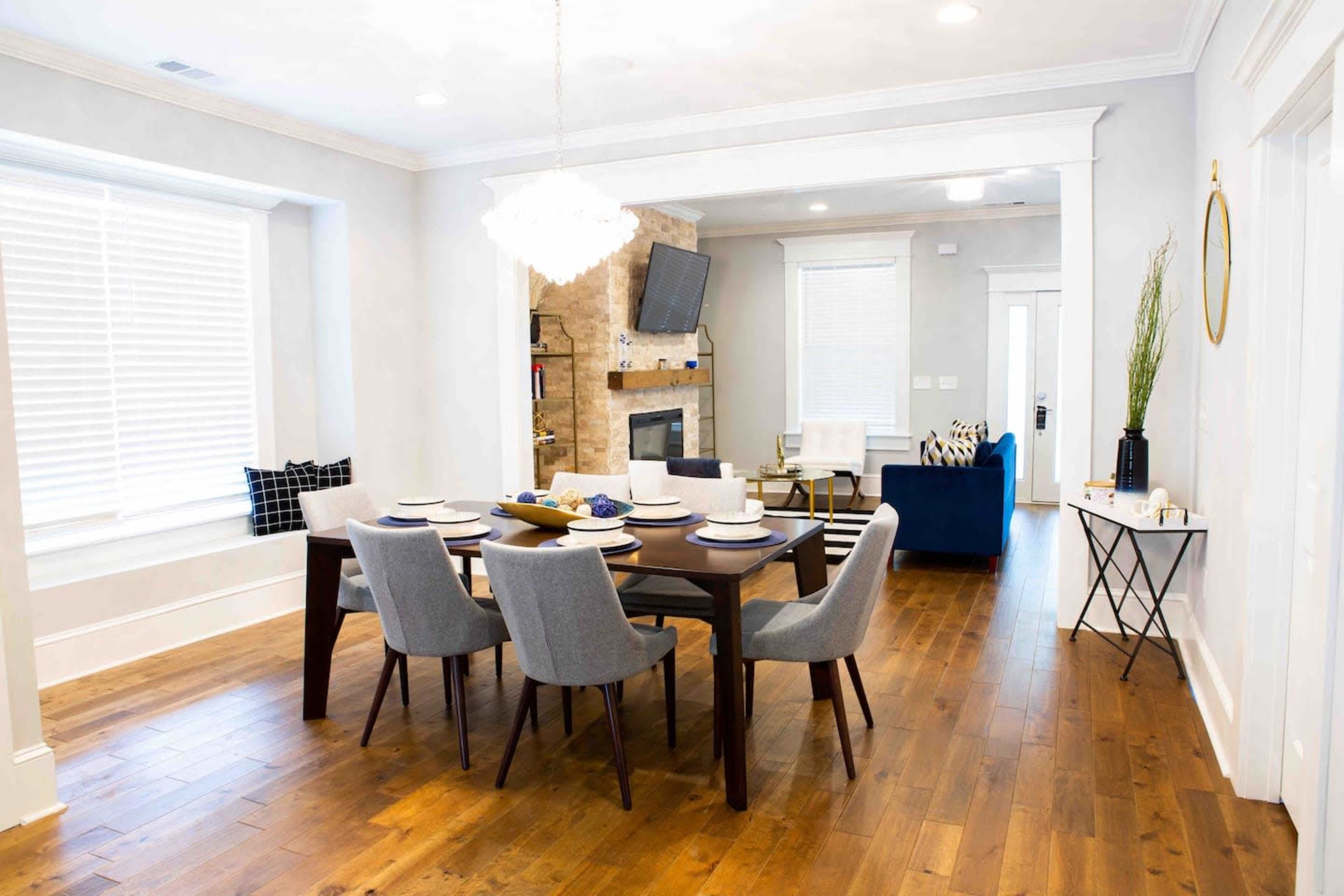 A dining area features a rectangular table set for eight, surrounded by gray chairs, with a stylish light fixture overhead and a cozy living room partially visible in the background.