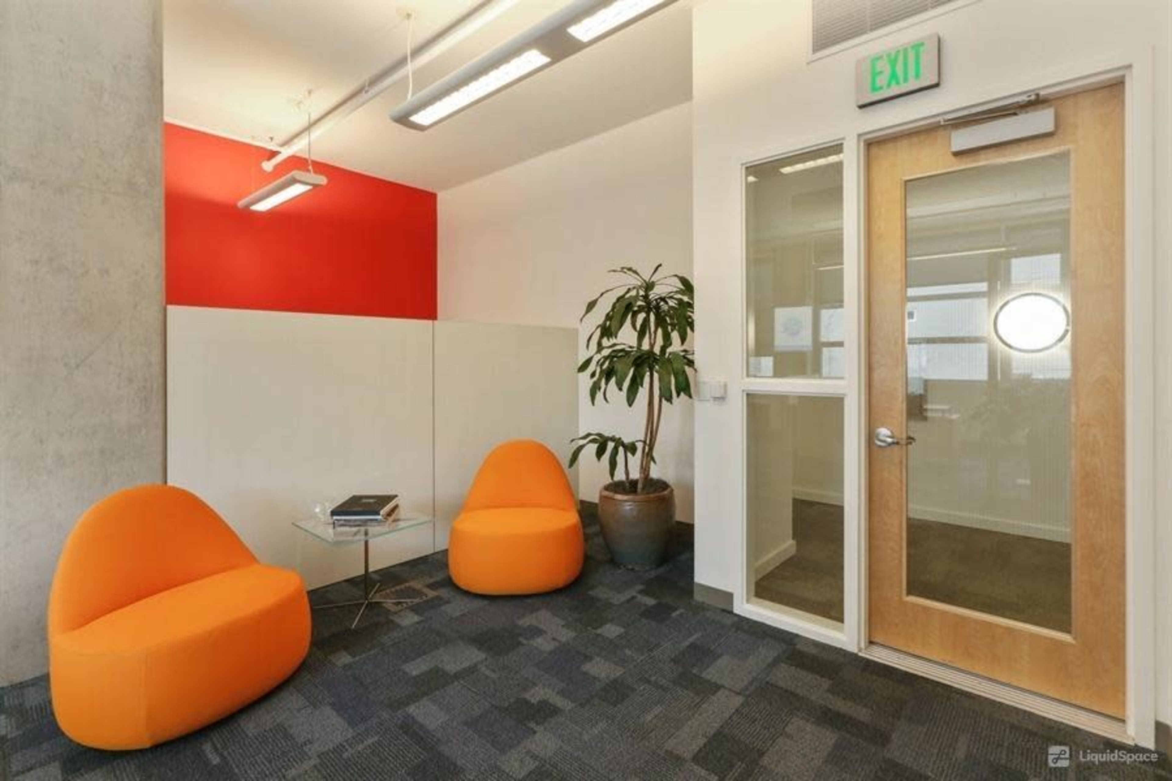 A small meeting area with two orange chairs, a glass coffee table, and a door leading to another room, set against a red accent wall.