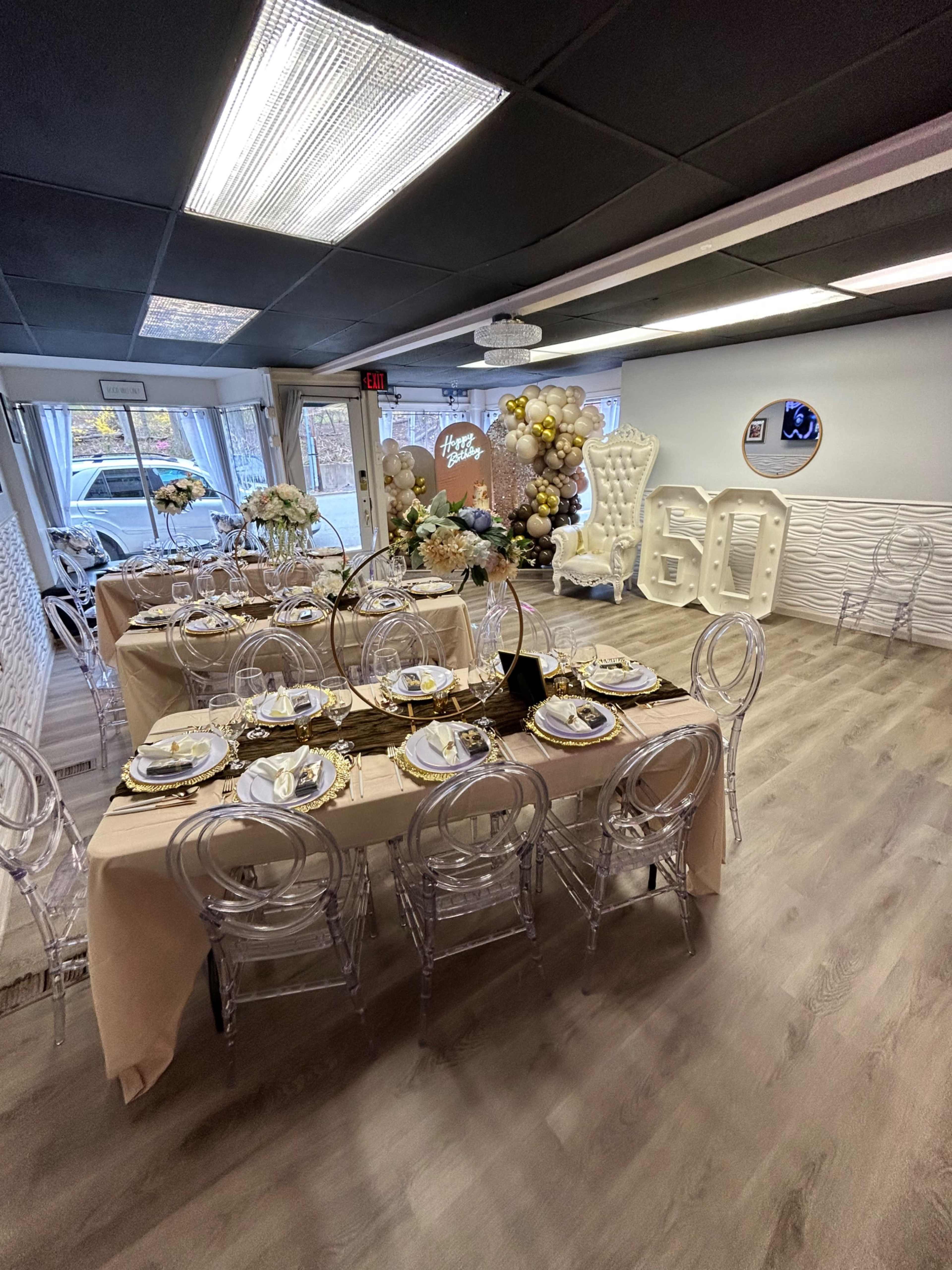 A decorated event space with elegantly set tables, clear chairs, and a backdrop featuring gold balloons and a "60" sign.
