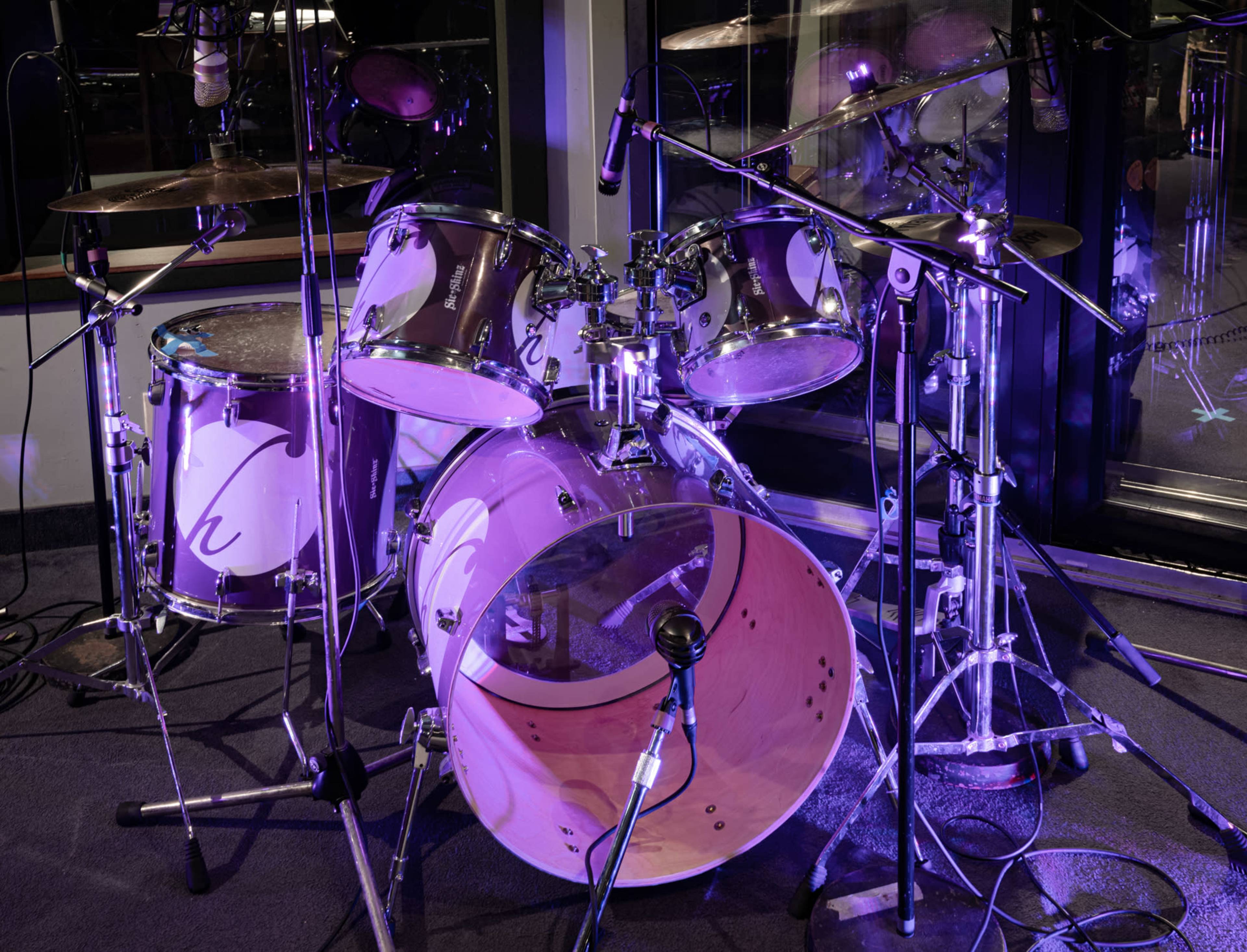A drum set with chrome hardware and transparent drumheads is set up in a recording studio, surrounded by microphones and soft lighting.