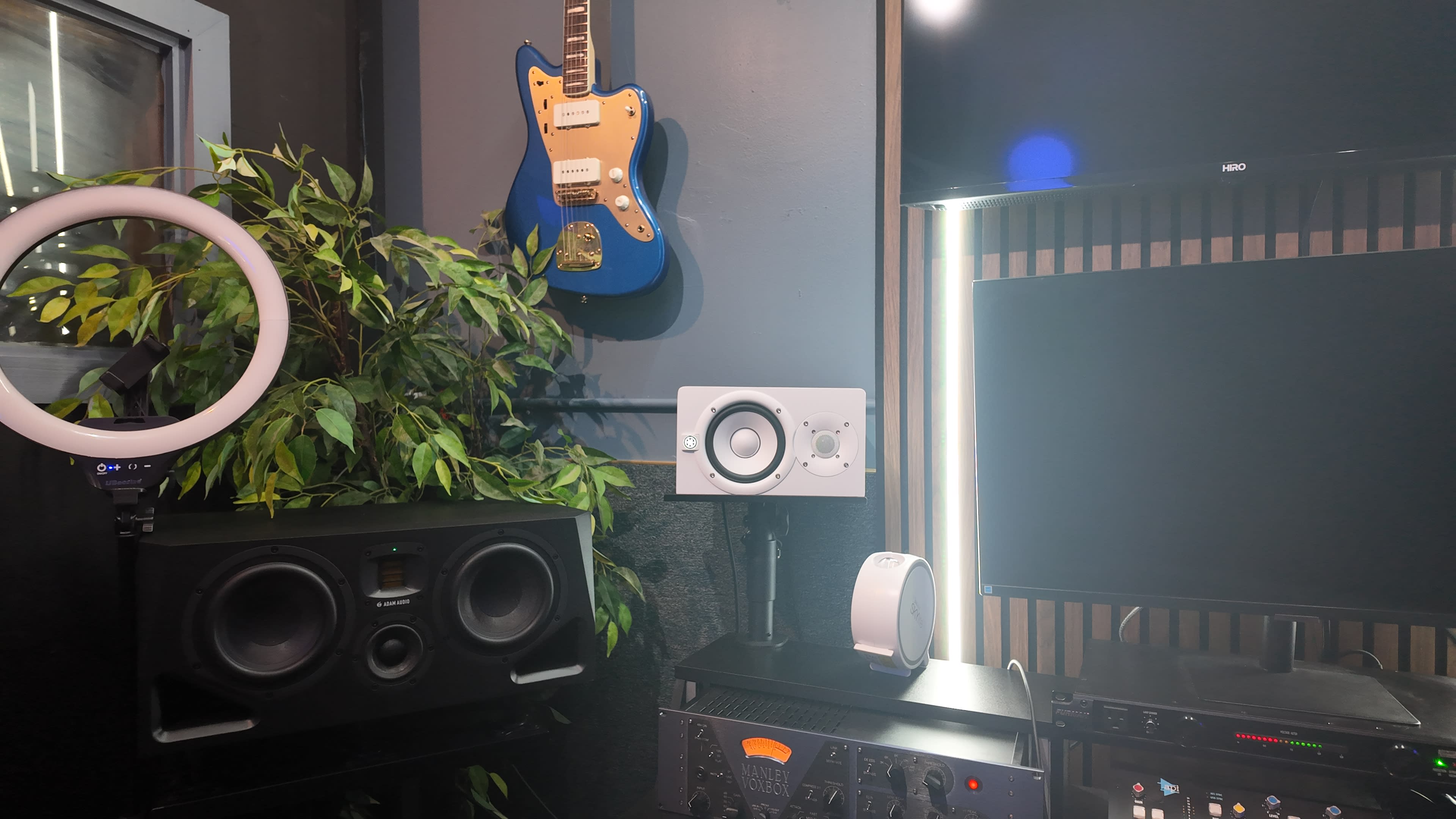 The image features a workspace with a blue electric guitar hanging on the wall, a potted plant, studio monitors, and a television screen.