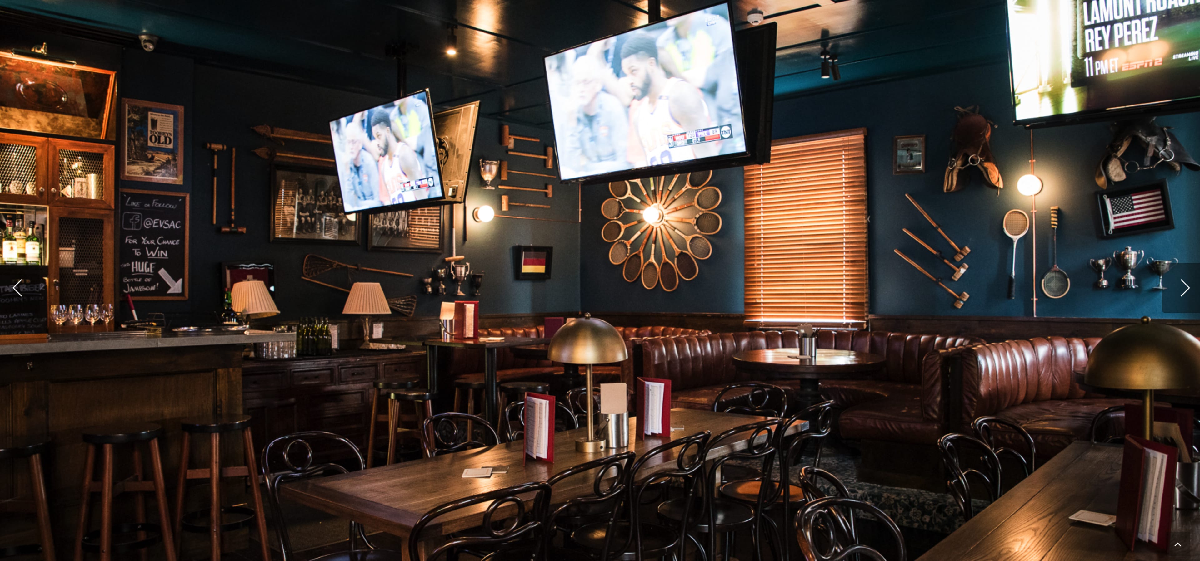 A cozy bar interior featuring leather seating, wooden tables, and two televisions displaying a sports game on the walls.