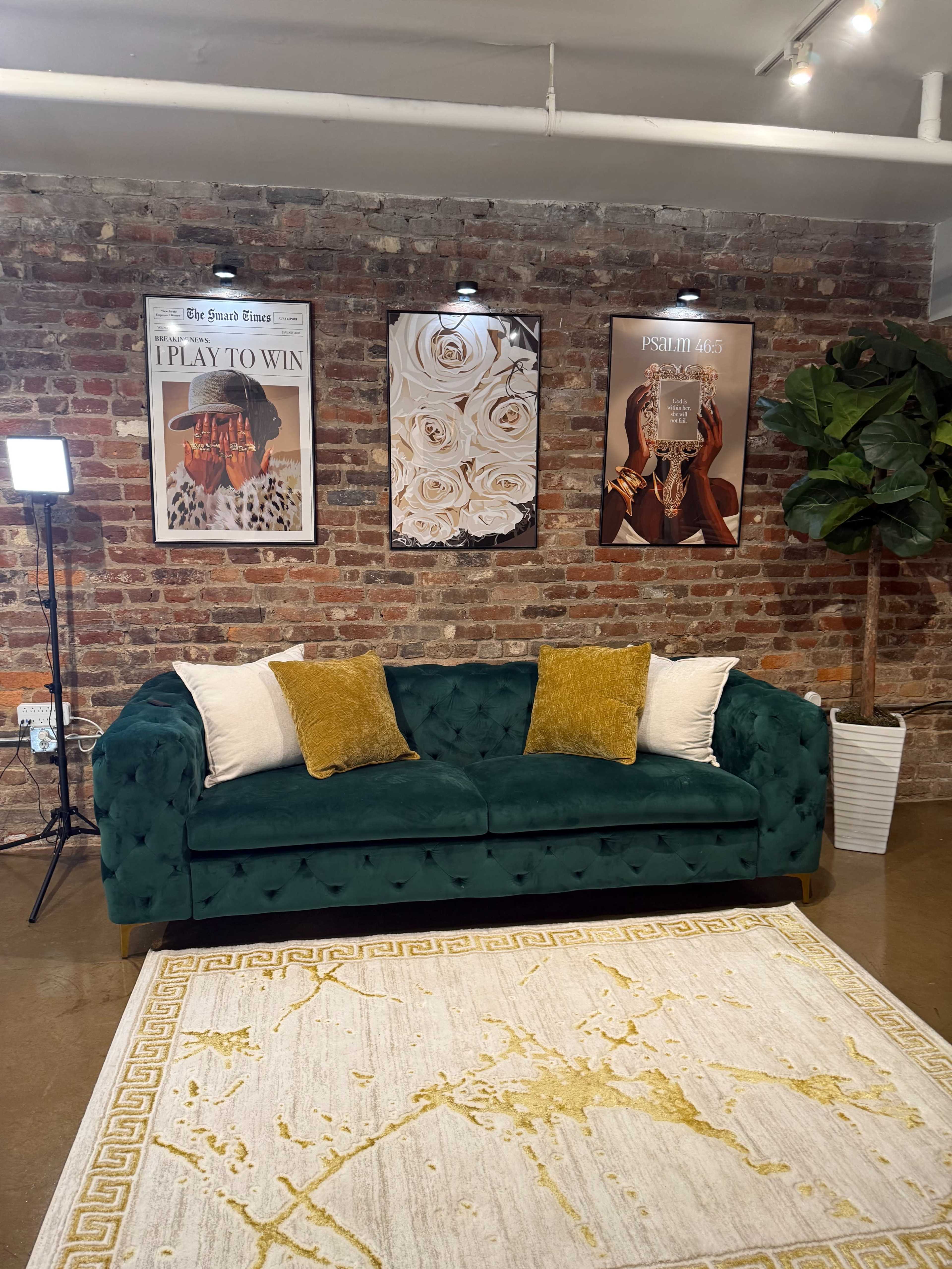 A green velvet couch with decorative pillows sits in front of a brick wall adorned with framed artwork and a tall potted plant nearby.