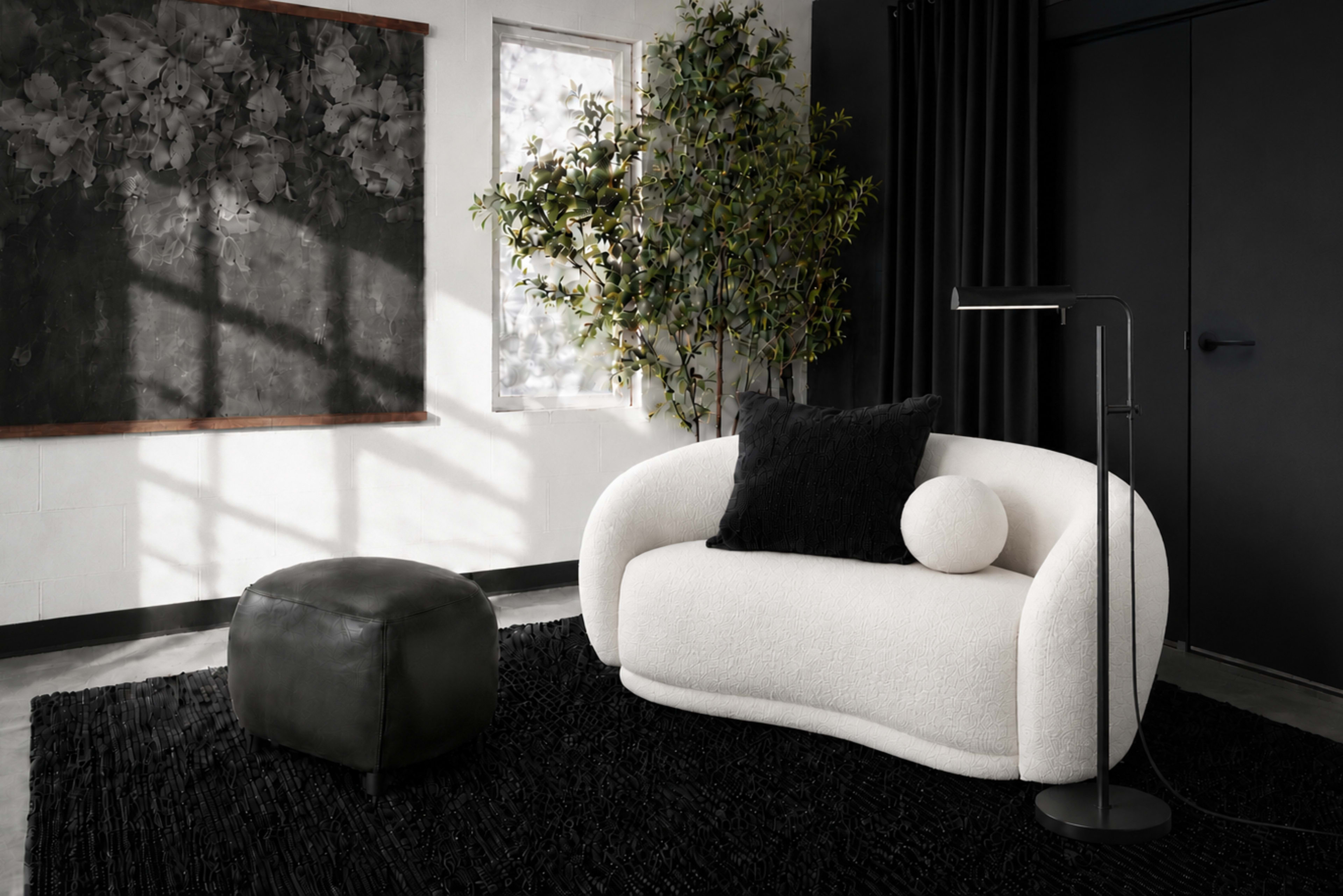 A curved white sofa with a black cushion and a round pillow sits beside a black ottoman on a black area rug in a room with large windows and a dark wall.