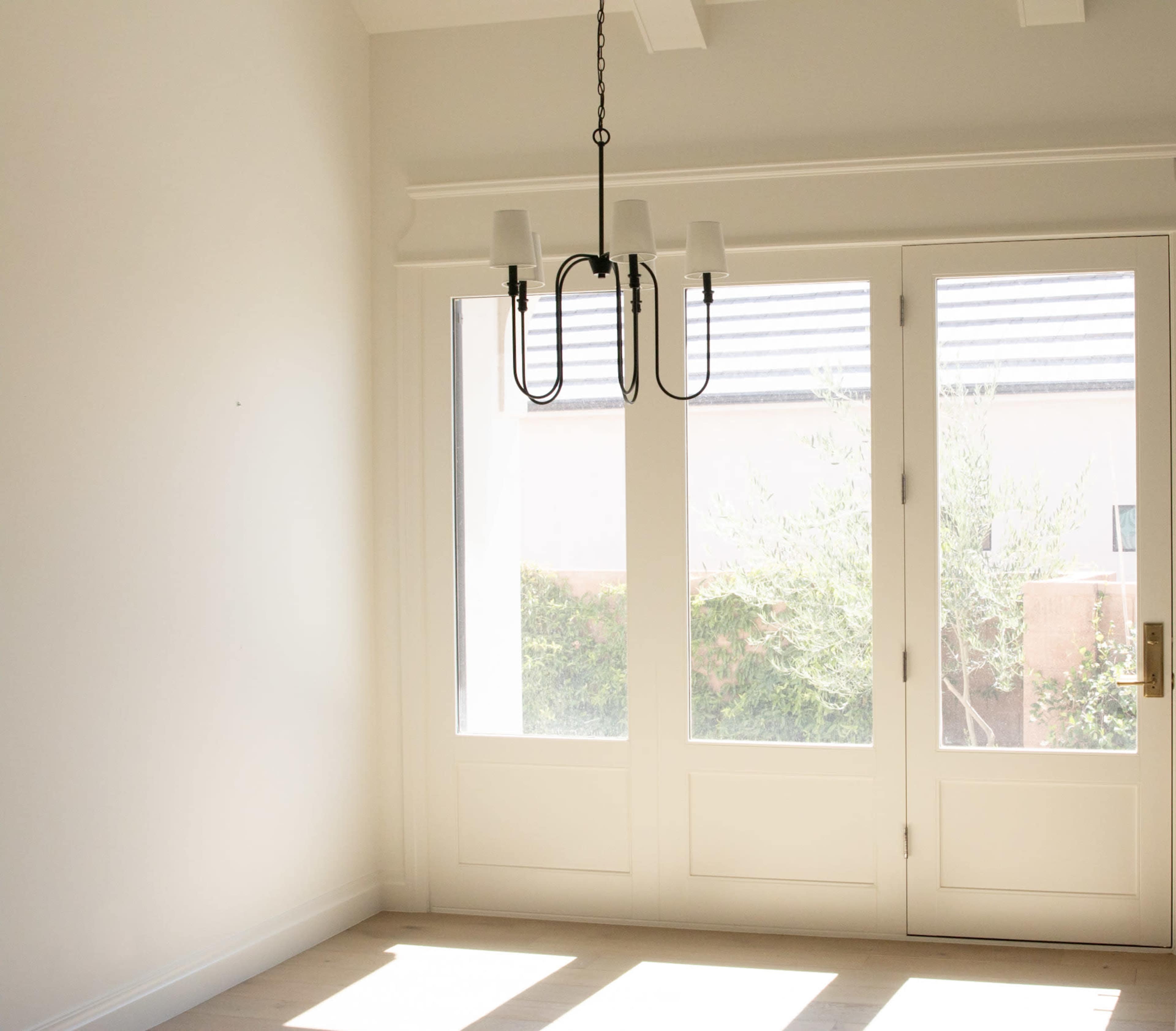 The image shows a bright, empty room with a chandelier hanging from the ceiling and large glass doors leading to an outdoor space.
