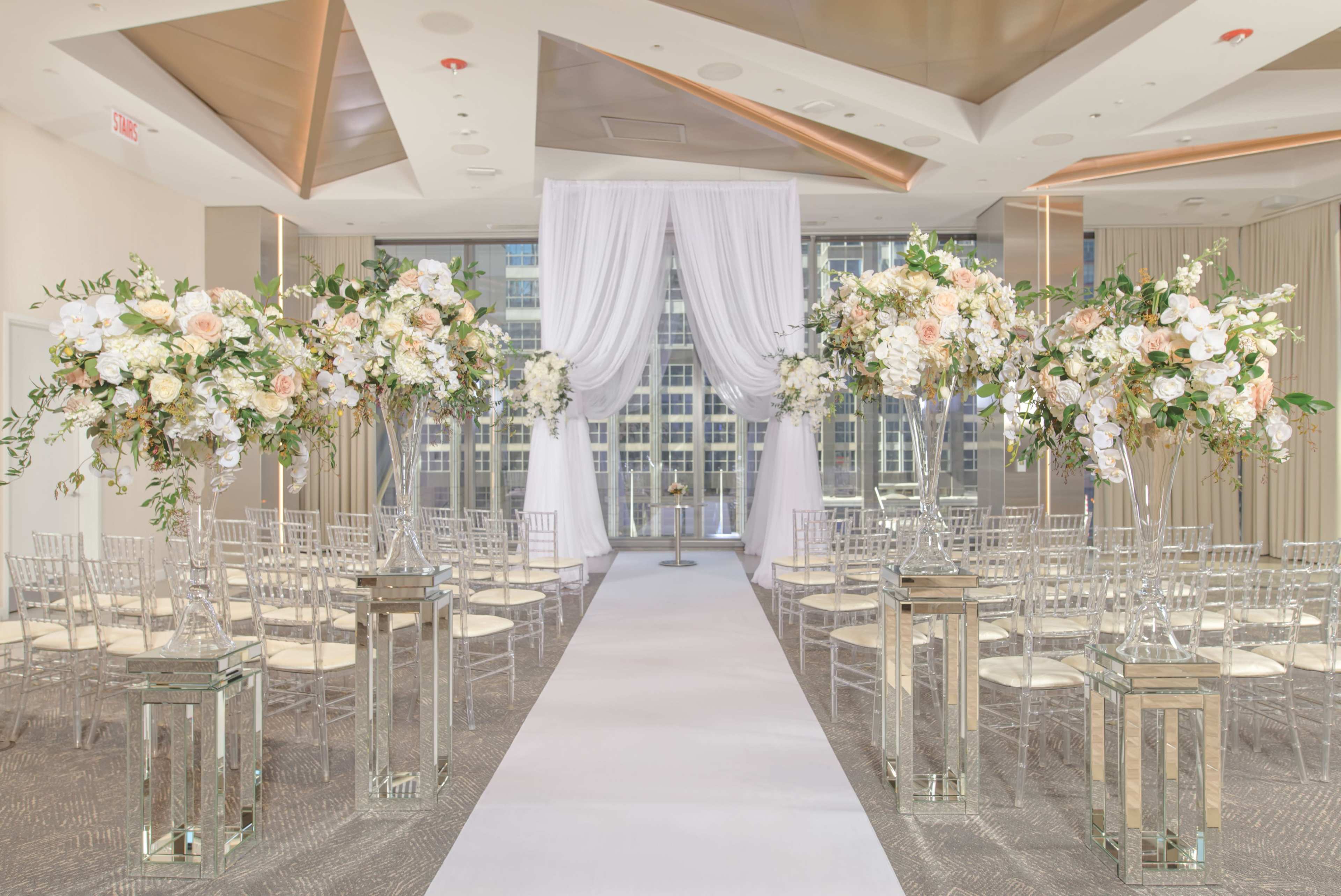 A wedding ceremony setup features rows of chairs aligned on a white aisle, flanked by tall floral arrangements and decorative lanterns, with sheer drapes framing a backdrop.