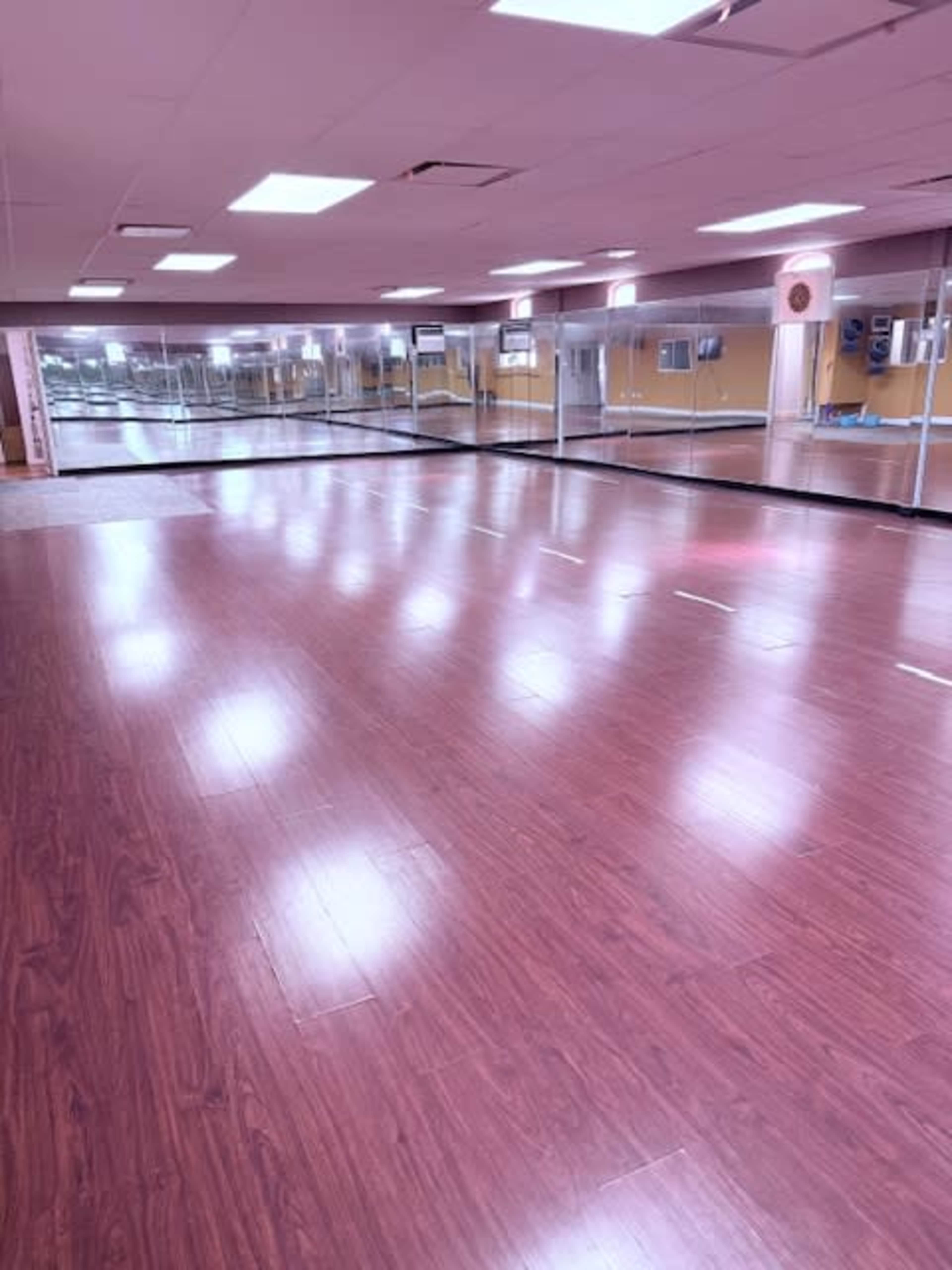 The image shows a spacious dance studio with wooden flooring and mirrored walls, illuminated by overhead lights.