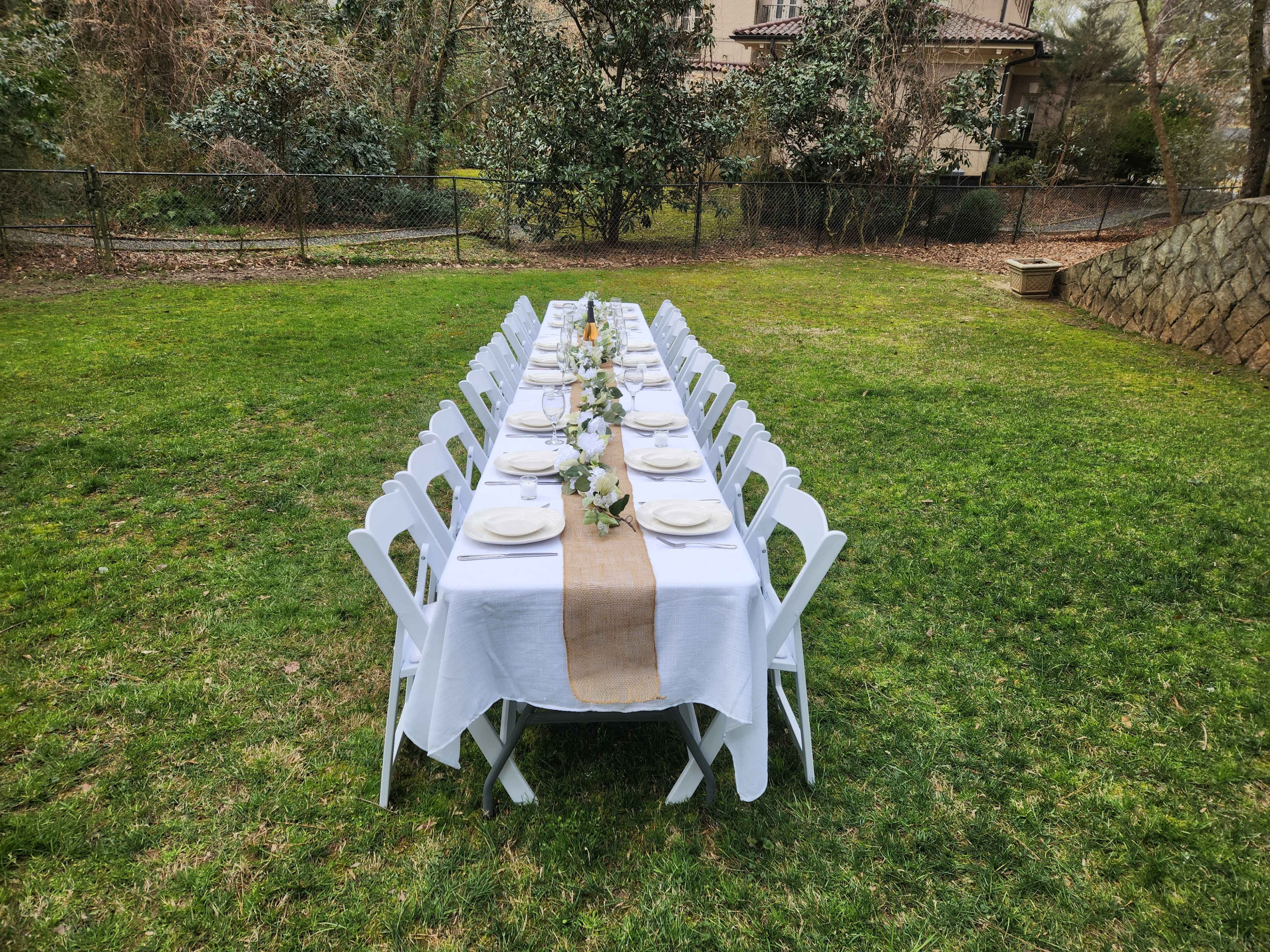 A long dining table is set with white tablecloths and plates, positioned on a grassy lawn surrounded by trees and a stone wall.