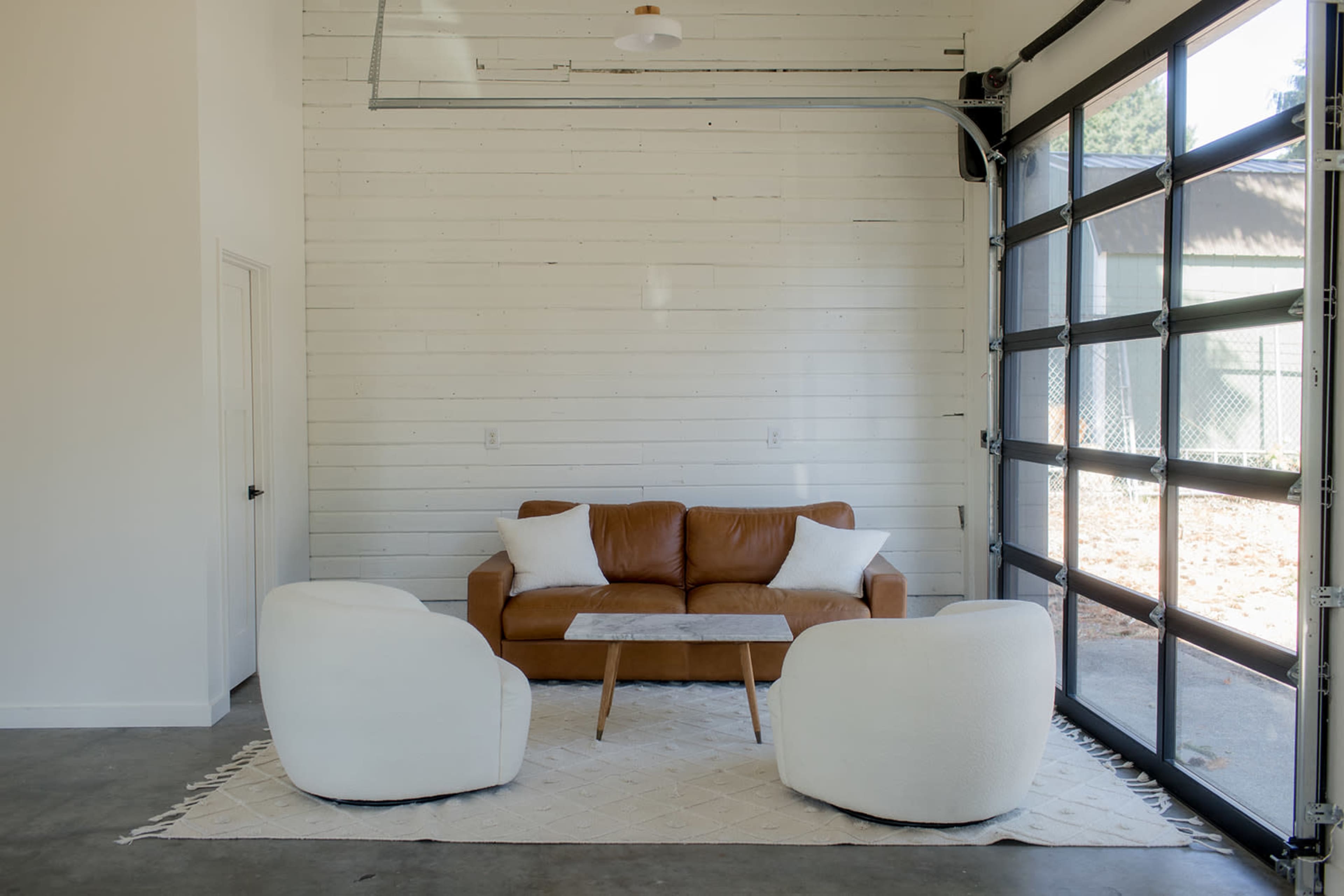 The image features a cozy seating area with a brown leather sofa, two white armchairs, and a small coffee table, set against a backdrop of a white wooden wall and a large glass garage door.