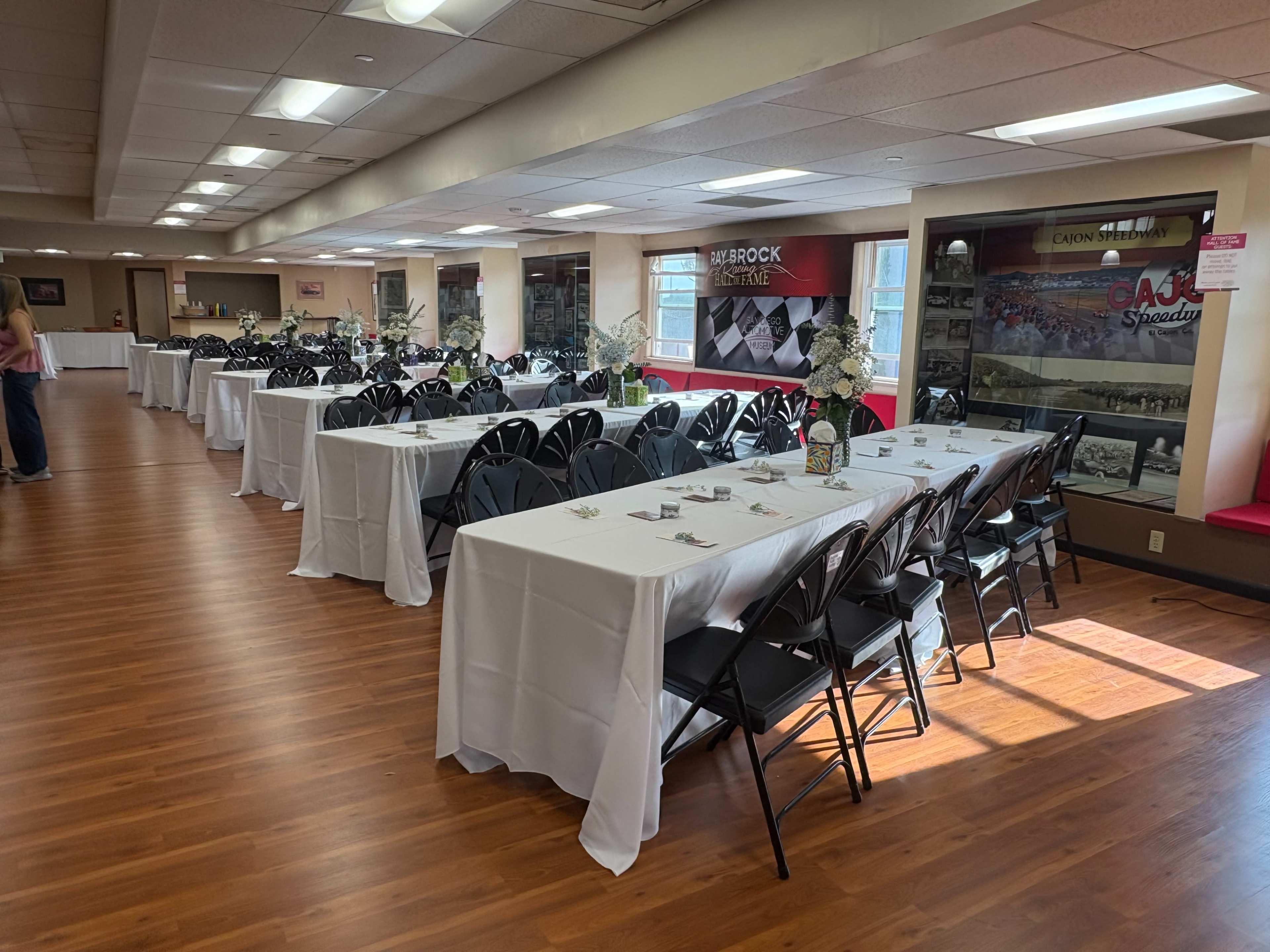 The image shows a banquet hall with multiple rows of tables covered in white tablecloths and arranged with black chairs, softly lit by natural light coming from windows on one side.