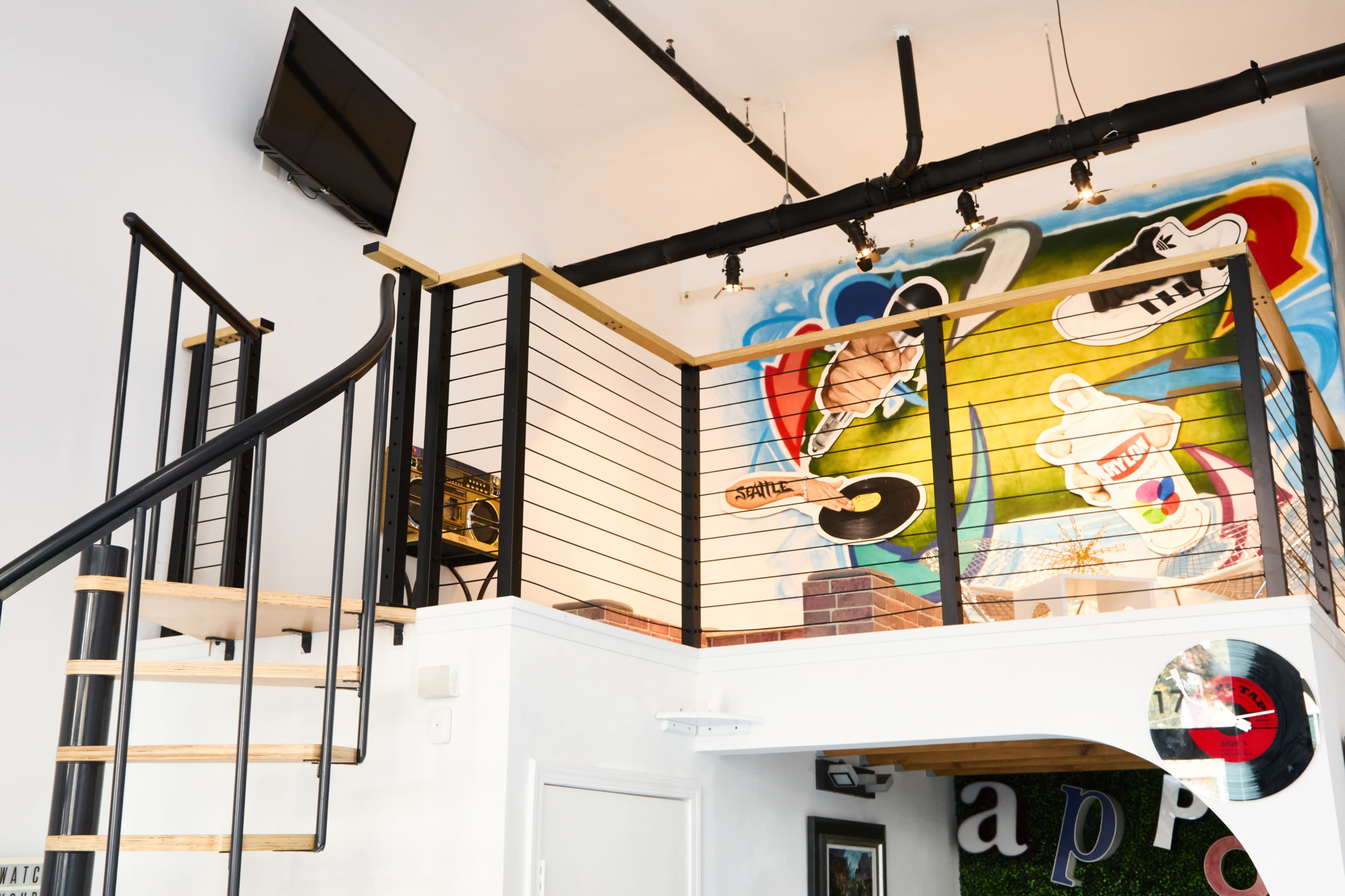 The image shows a modern interior with a spiral staircase leading to an upper level, featuring a colorful mural and a wall-mounted television.