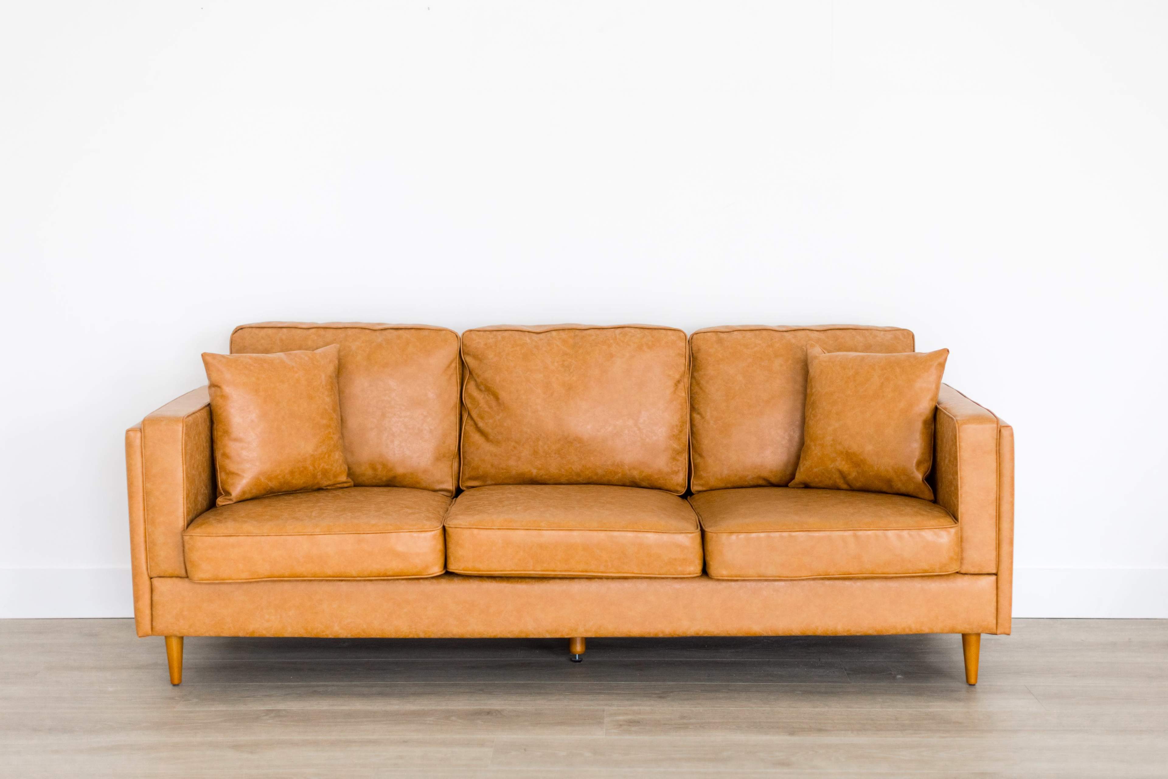 A tan leather sofa with three seat cushions and two matching pillows is positioned against a white wall.
