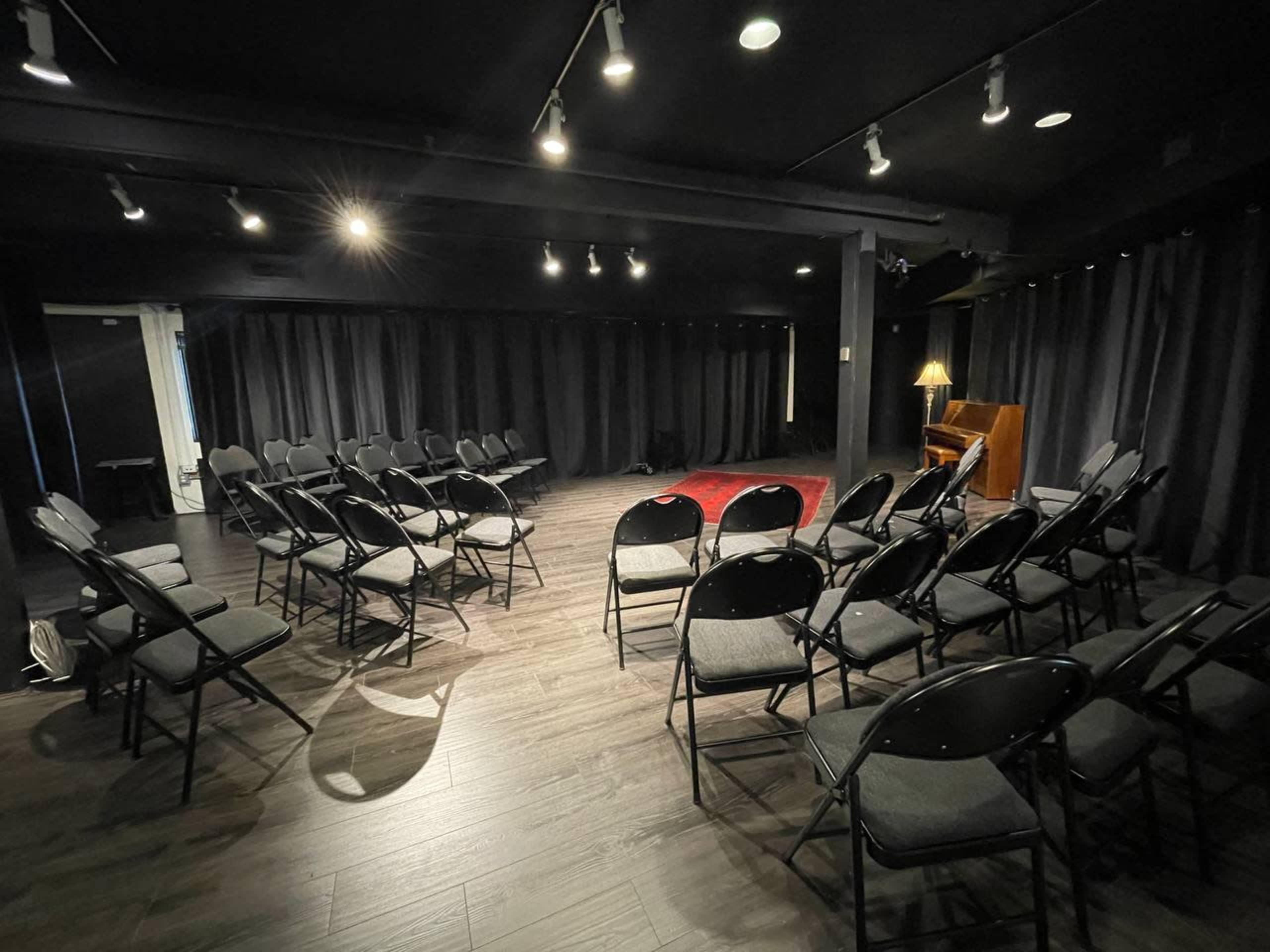 A dimly lit performance space with a circle of black chairs arranged on a wooden floor, accompanied by a small piano and a lamp in the background.
