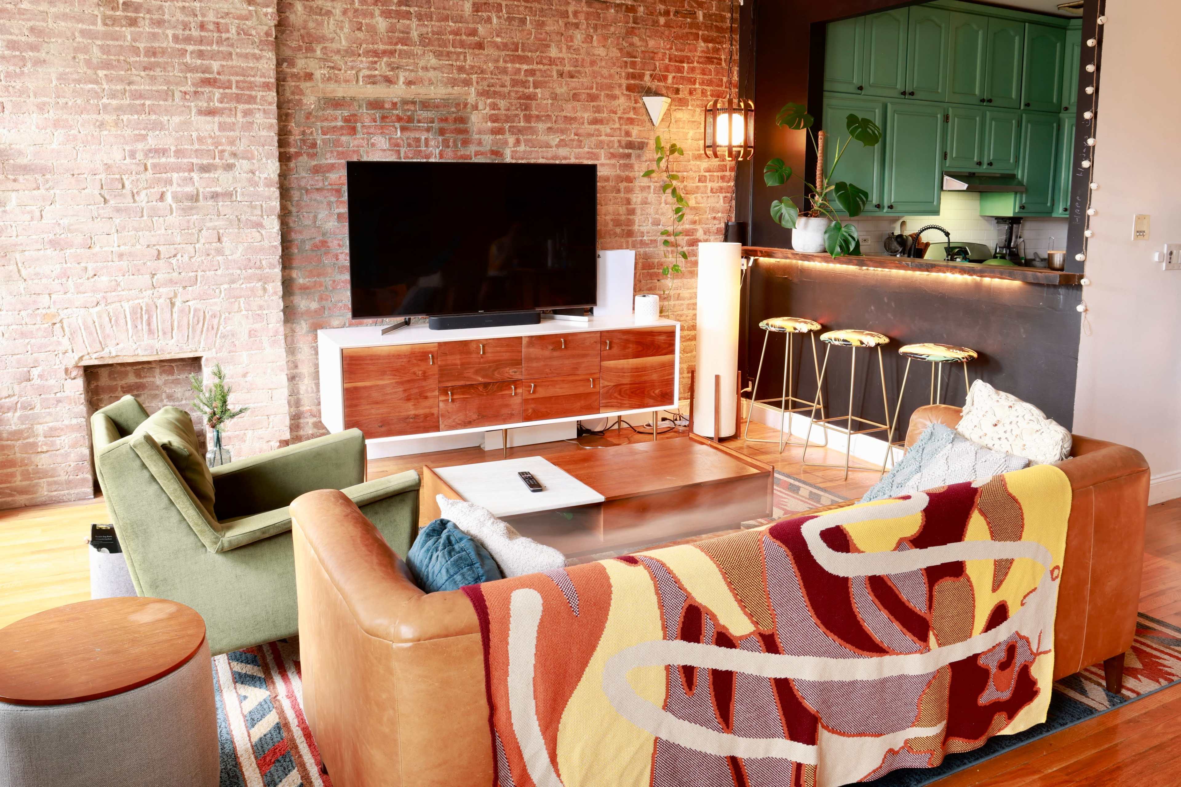 The image shows a cozy living room with exposed brick walls, a large television, and a kitchen area featuring green cabinets and bar stools.