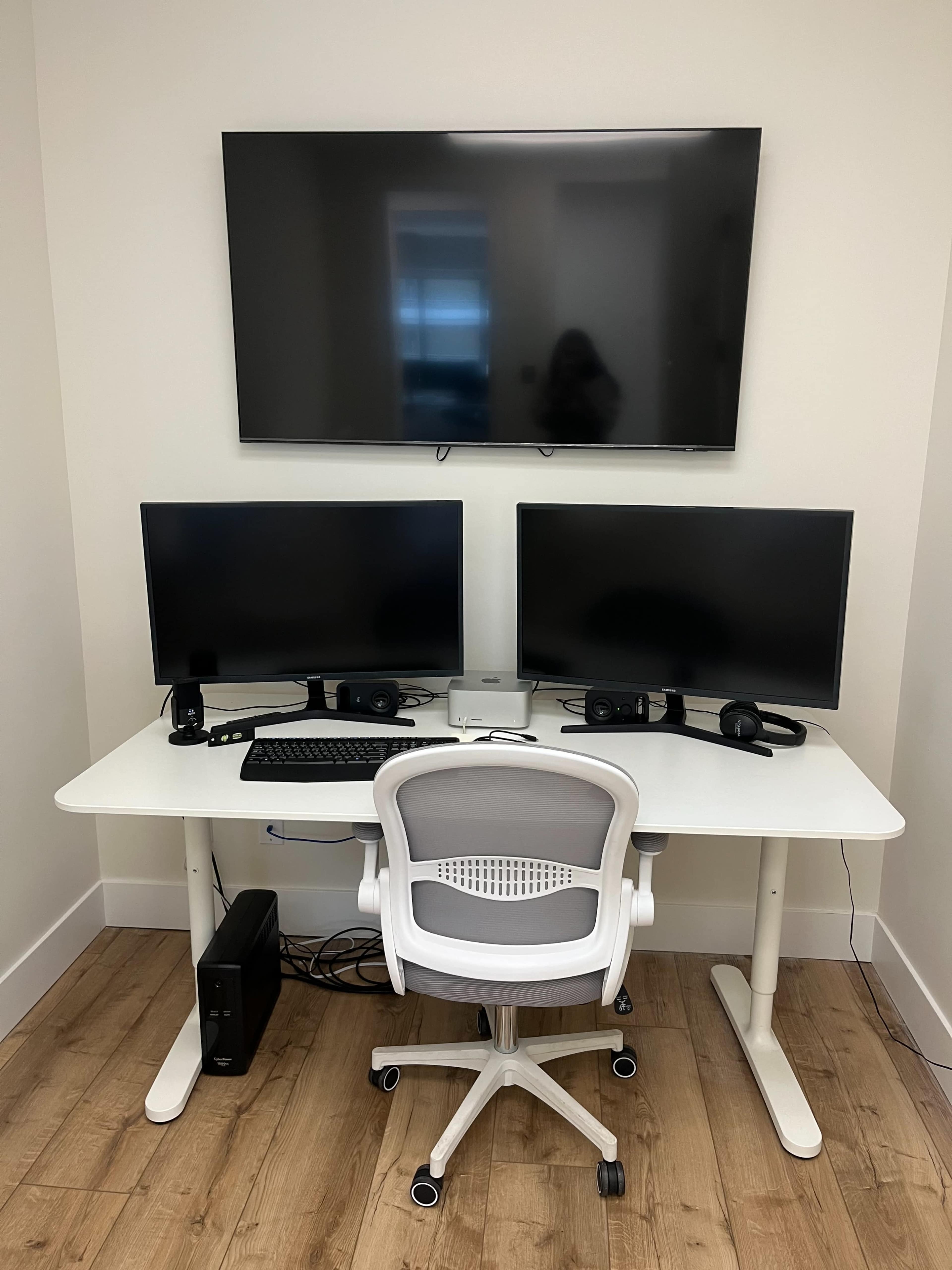 The image shows a modern workstation with two computer monitors, a keyboard, a mouse, a headset, and a wall-mounted TV above a white desk, accompanied by a rolling office chair.