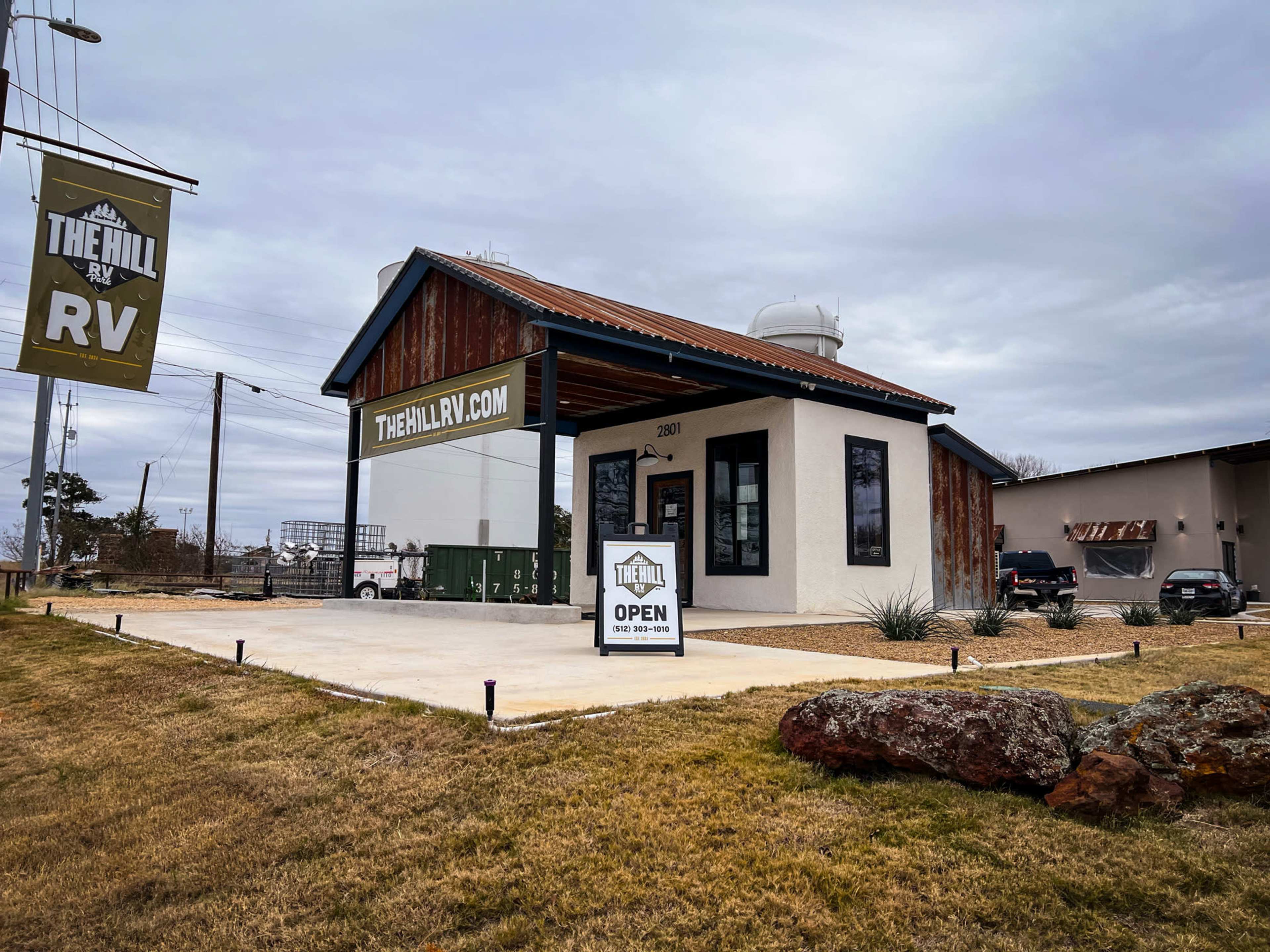 A modern building with a sloped roof features a sign reading "THEHILLRV.COM" and an "OPEN" sign on the front, surrounded by a parking lot and landscaping.