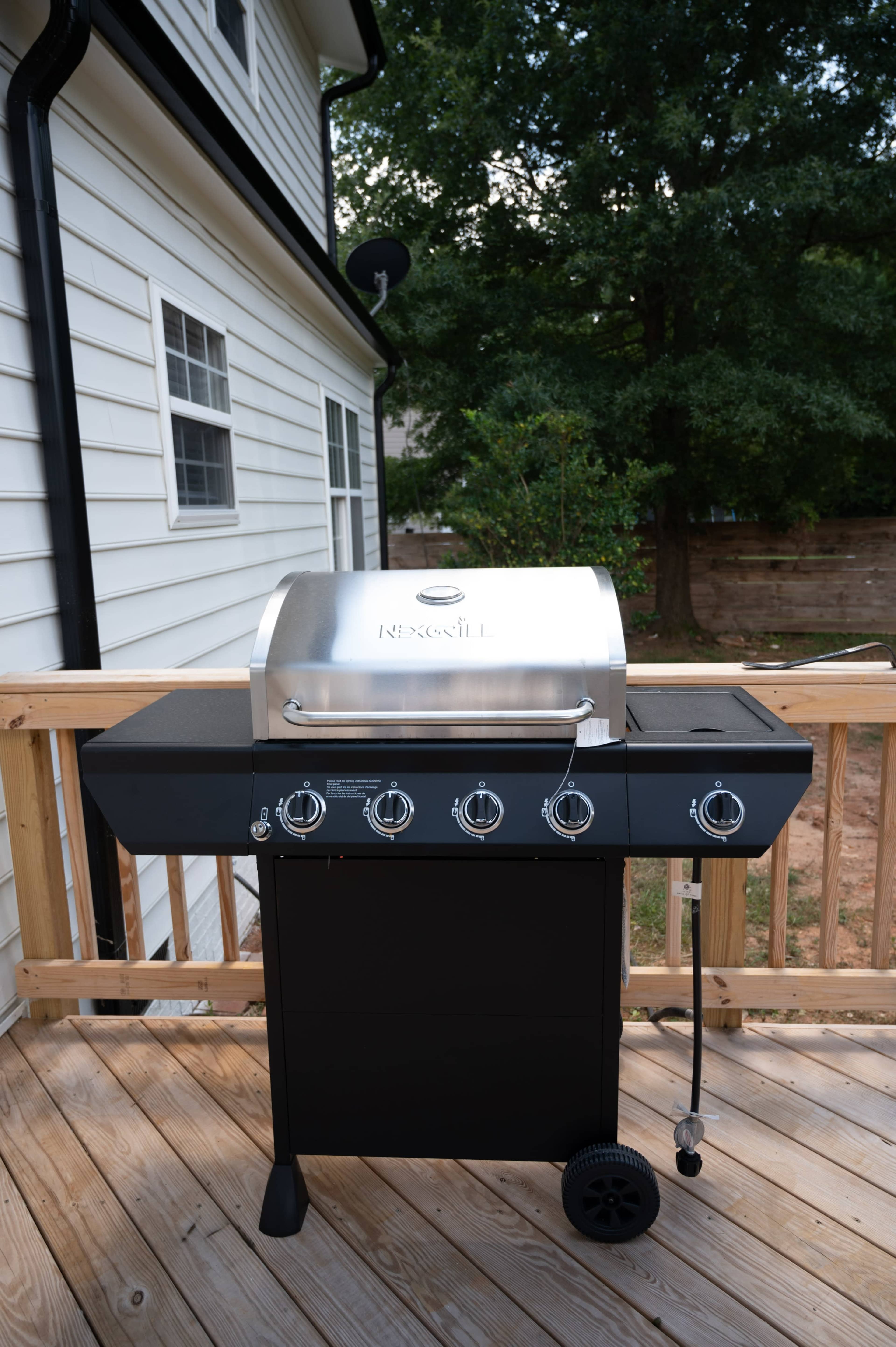 The image shows a stainless steel gas grill positioned on a wooden deck next to a white house.