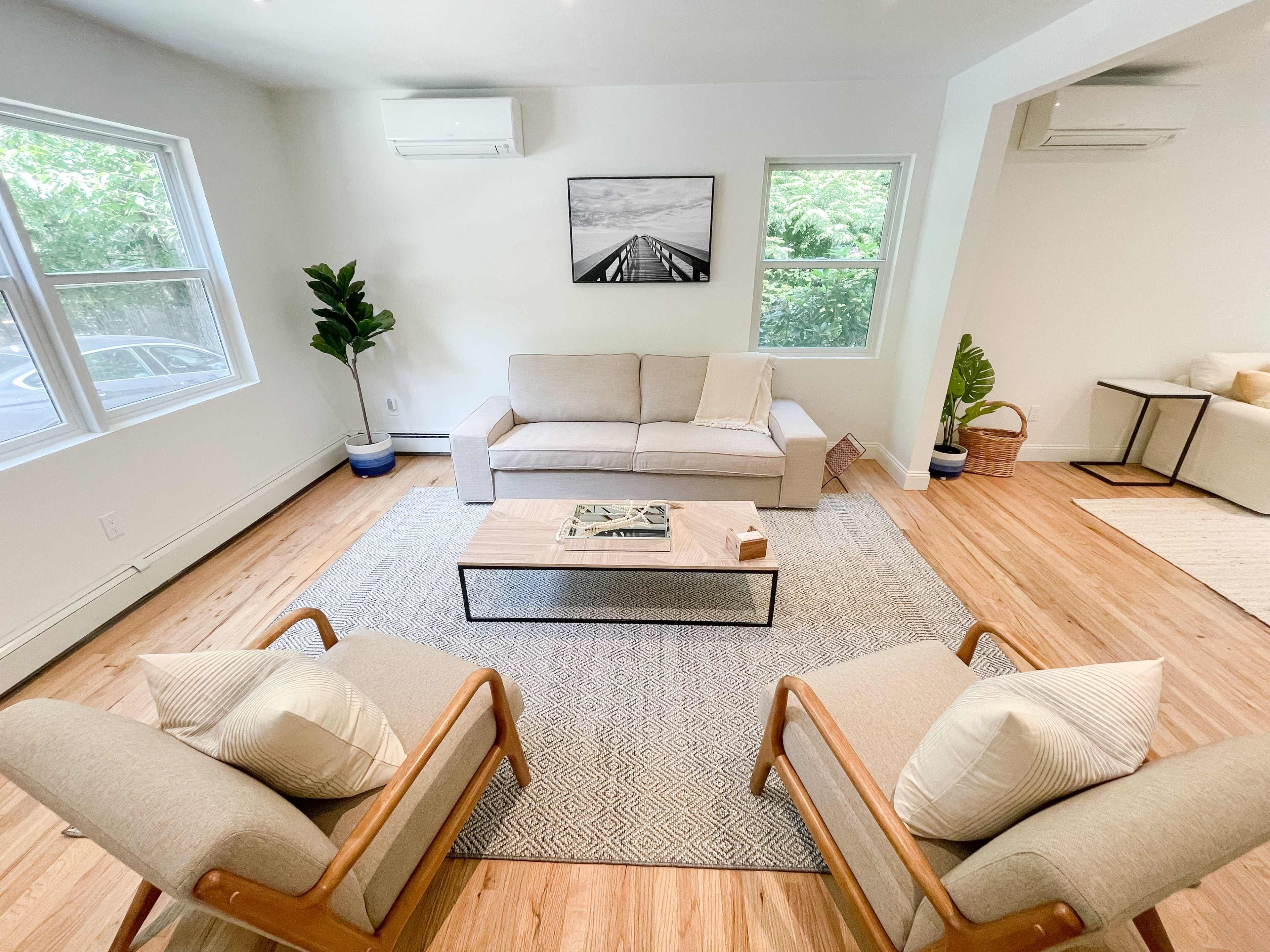 A bright living room with a light-colored sofa, two wooden armchairs, a coffee table in the center, and large windows allowing natural light to enter.