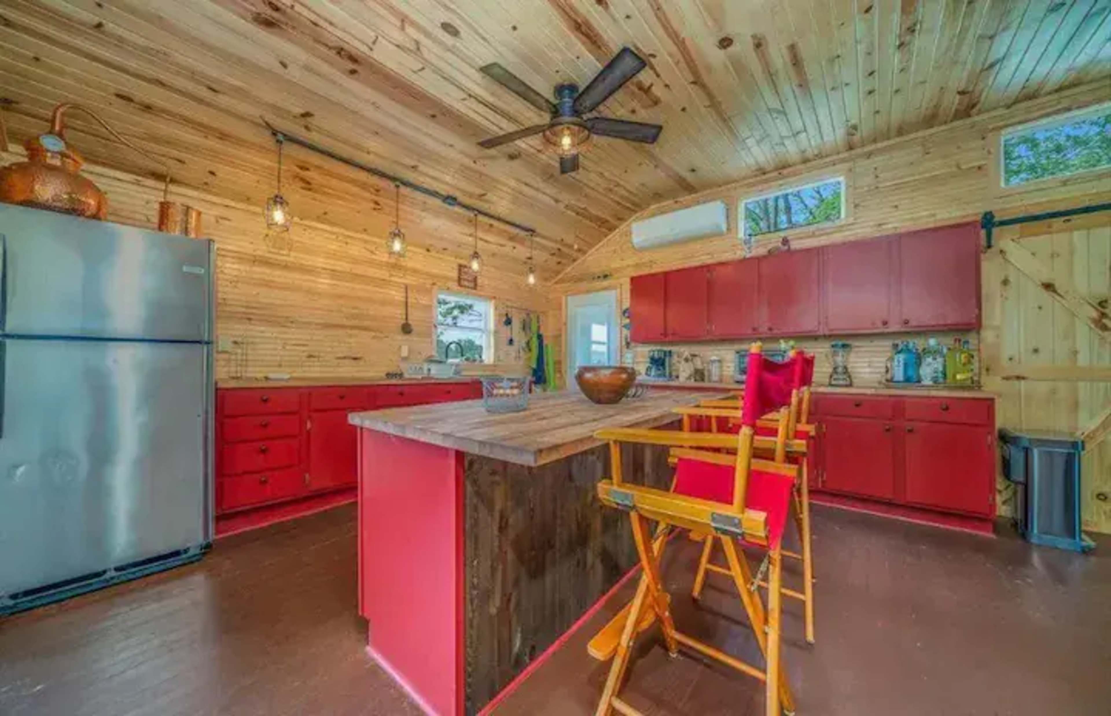 A rustic kitchen with wooden walls and ceiling, featuring red cabinets, stainless steel appliances, and a wooden island with tall chairs.