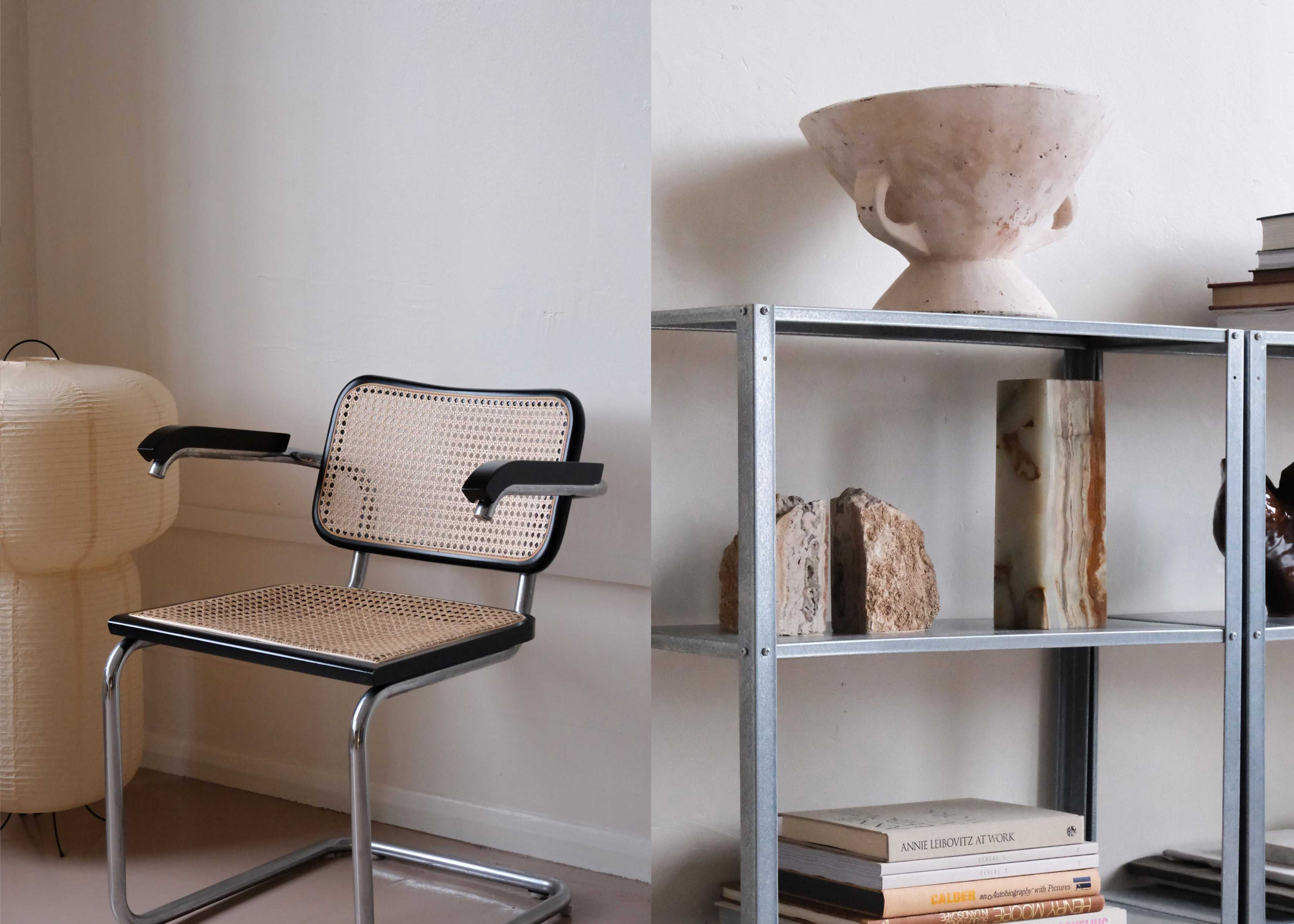 A modern room featuring a metal-framed chair with a woven seat alongside a decorative bowl atop a metal shelf filled with books and various objects.