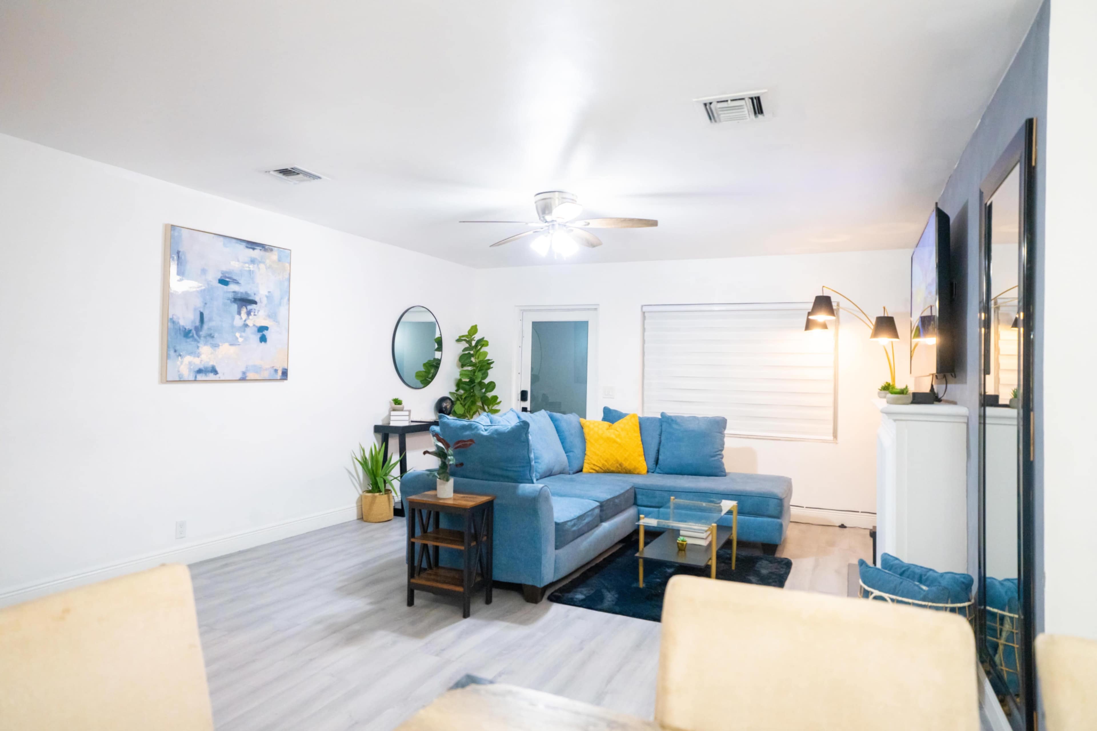 The image shows a modern living room featuring a blue sectional sofa with yellow accent pillows, a round mirror, and a small table by the window.