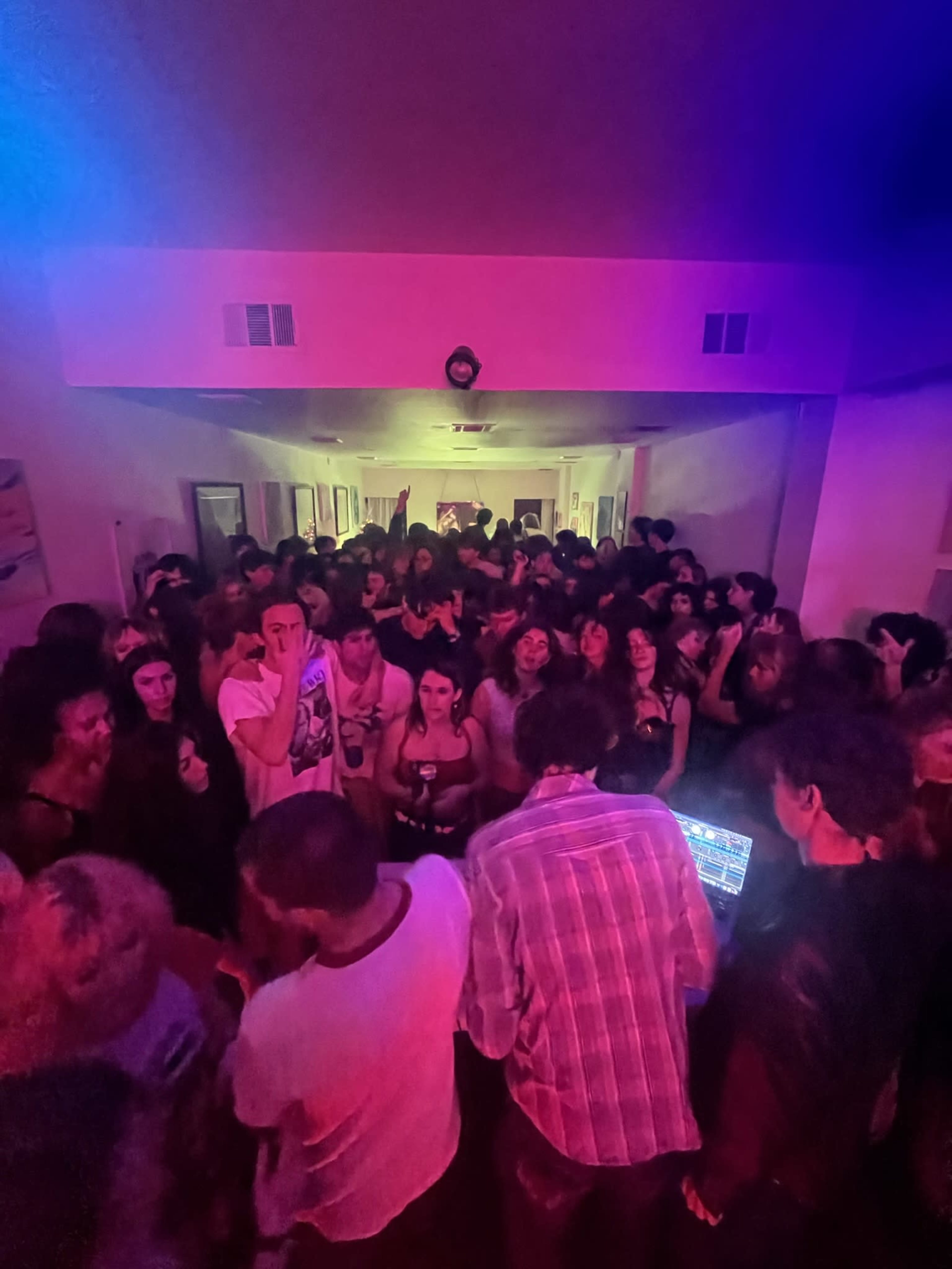 A crowded party scene is captured in a dimly lit hallway with vivid colored lights and a DJ setup at the forefront.
