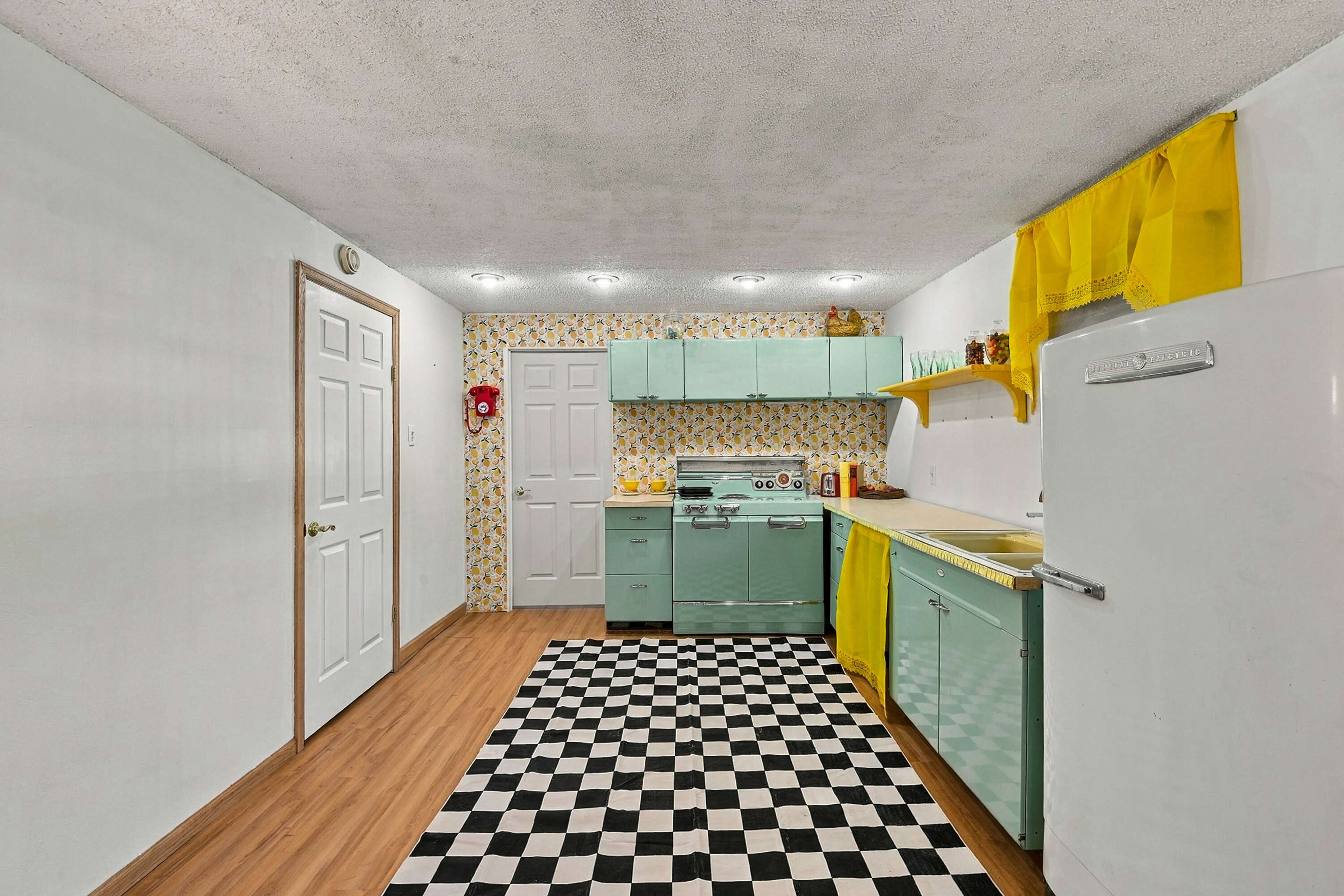 The image shows a vintage kitchen with mint green cabinets, a white fridge, a checkerboard black and white floor, and yellow accents.