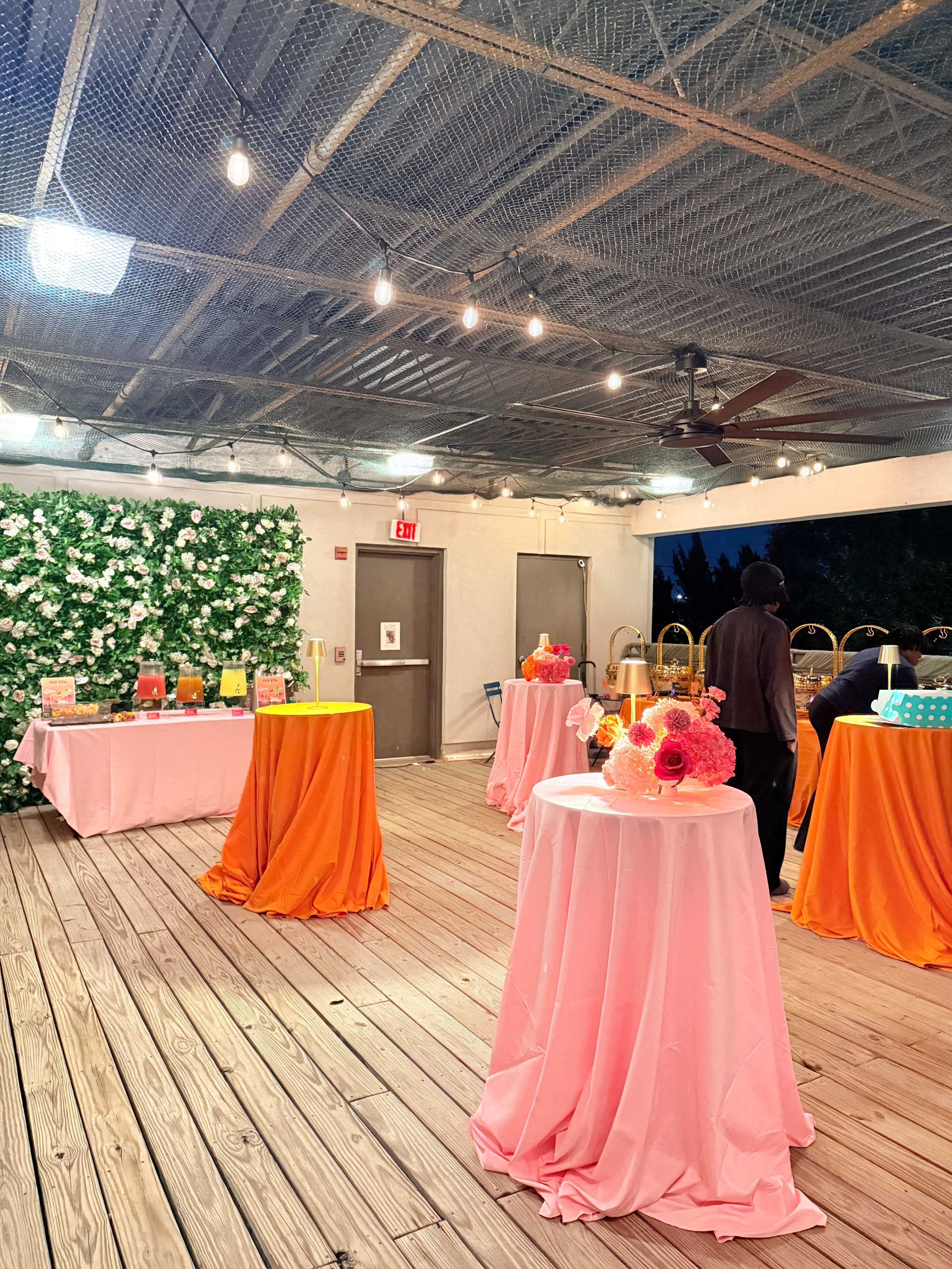 The scene features a decorated indoor space with tables draped in pink and orange cloths, surrounded by greenery and string lights.