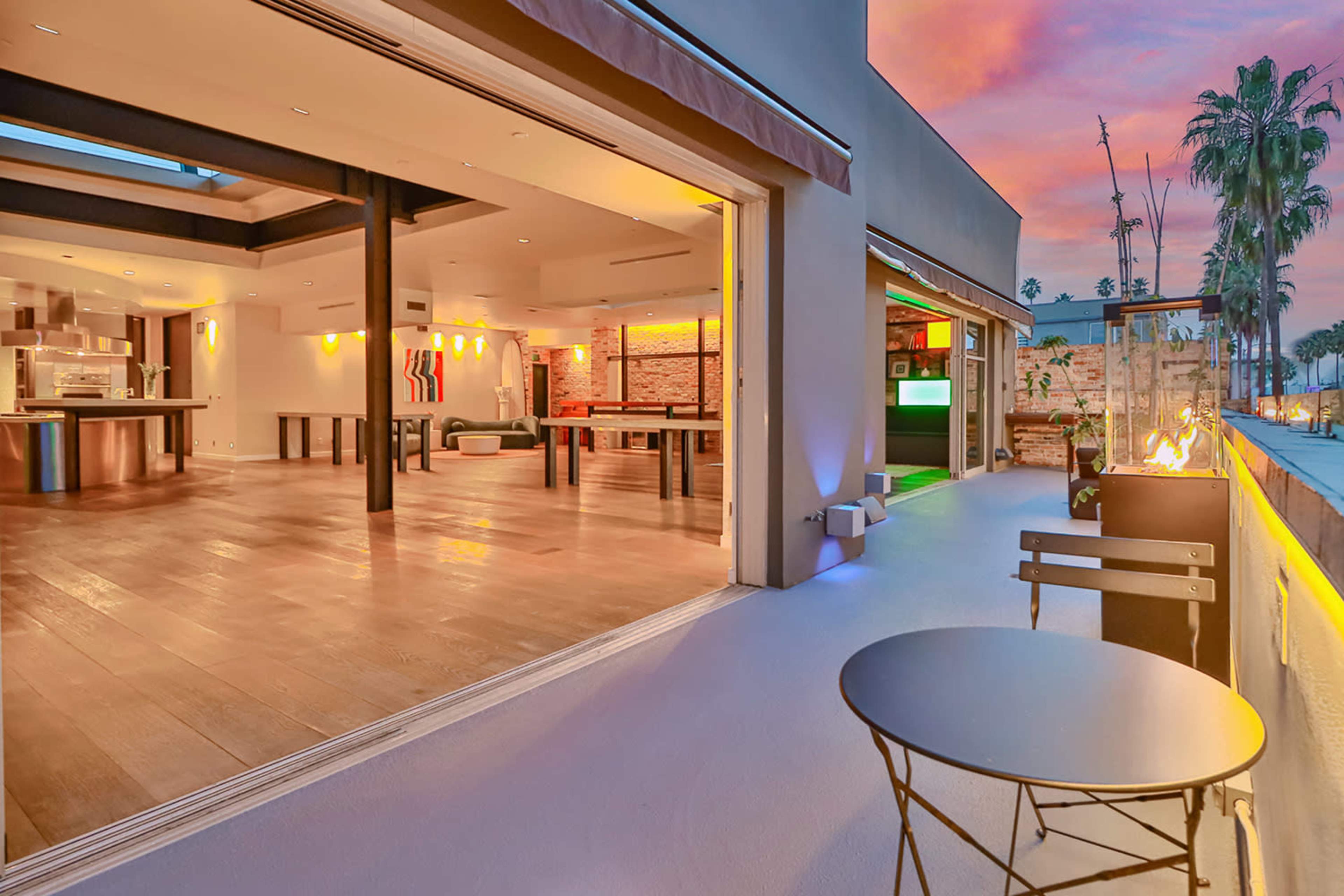 A modern outdoor space features an open-concept living area with a kitchen and seating, illuminated by ambient lighting against a colorful sunset backdrop.