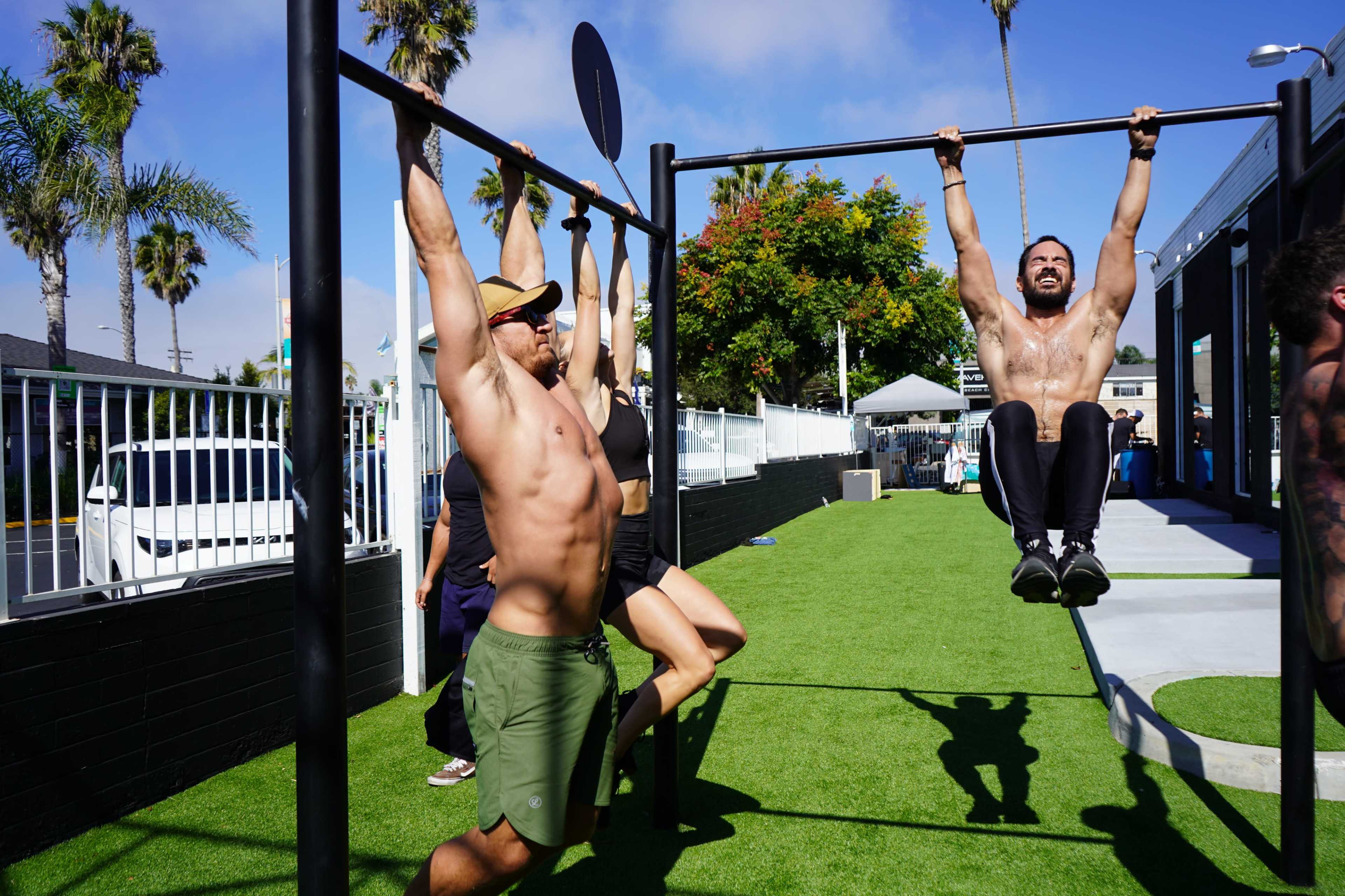 Two men perform pull-ups on a bar while a woman exercises nearby in a sunny outdoor gym.